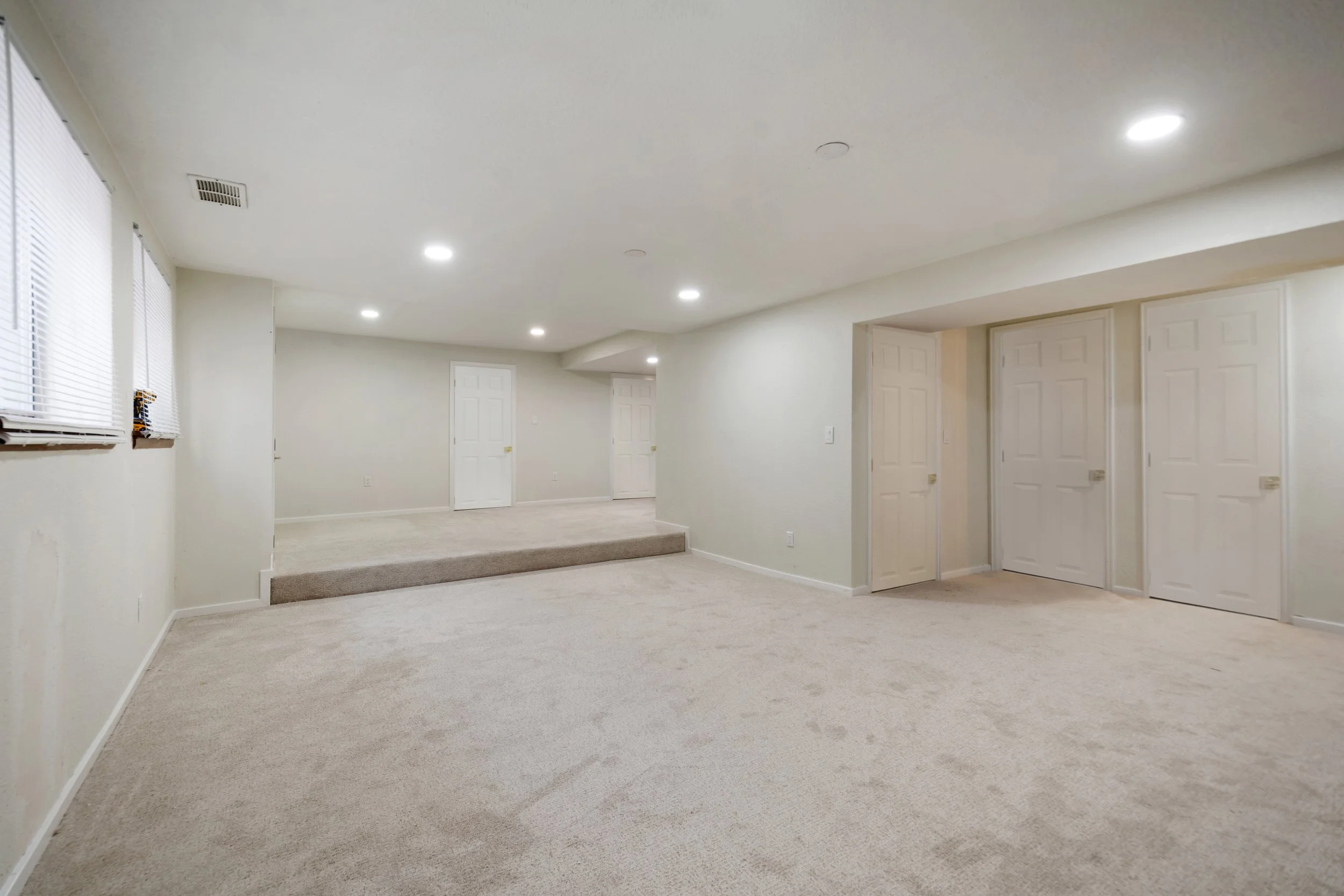 Empty beige basement room with carpeted floor, white walls, multiple white closets, and ceiling lights