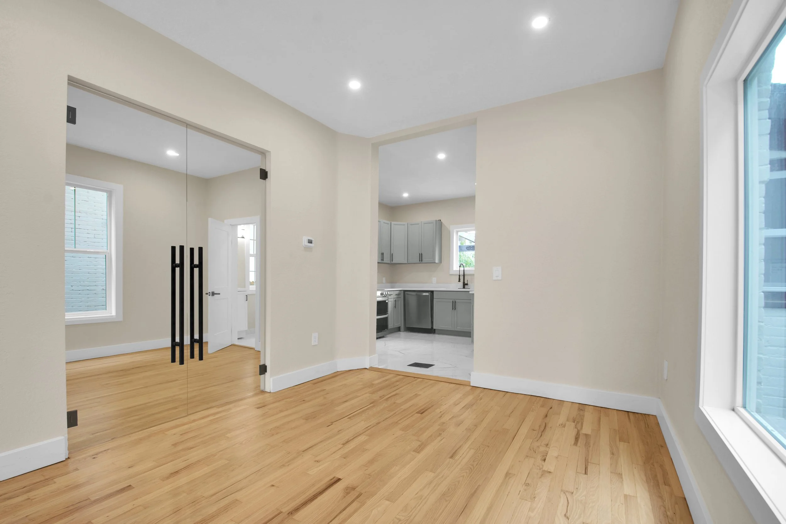 Empty living room with light-colored walls, hardwood floors, large window, and glass double doors leading to another room with white trim.