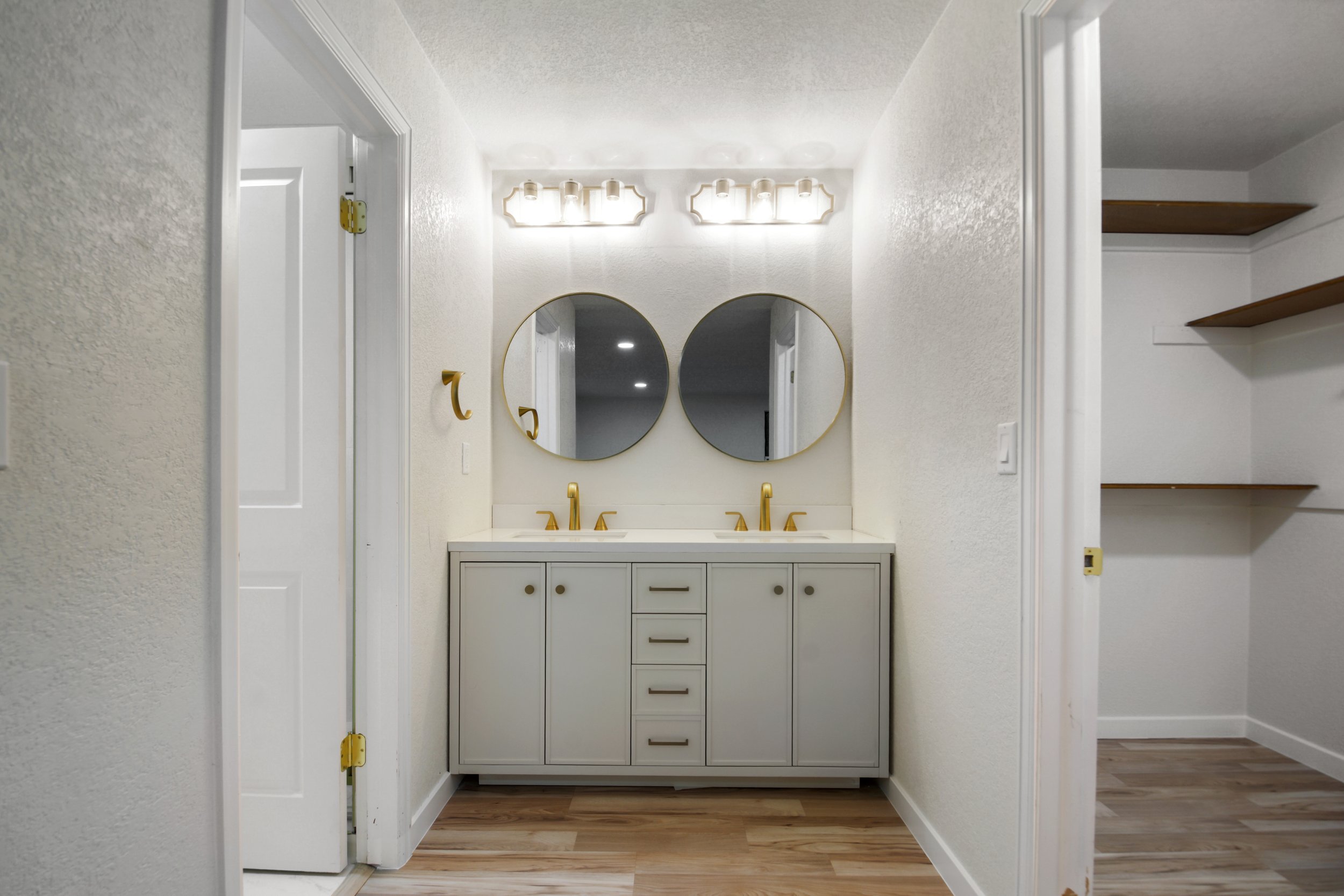 Bathroom vanity with two sinks, two round mirrors with gold frames, and gold fixtures, illuminated by a row of vanity lights, with an open shelving closet to the right and closed doors on either side.