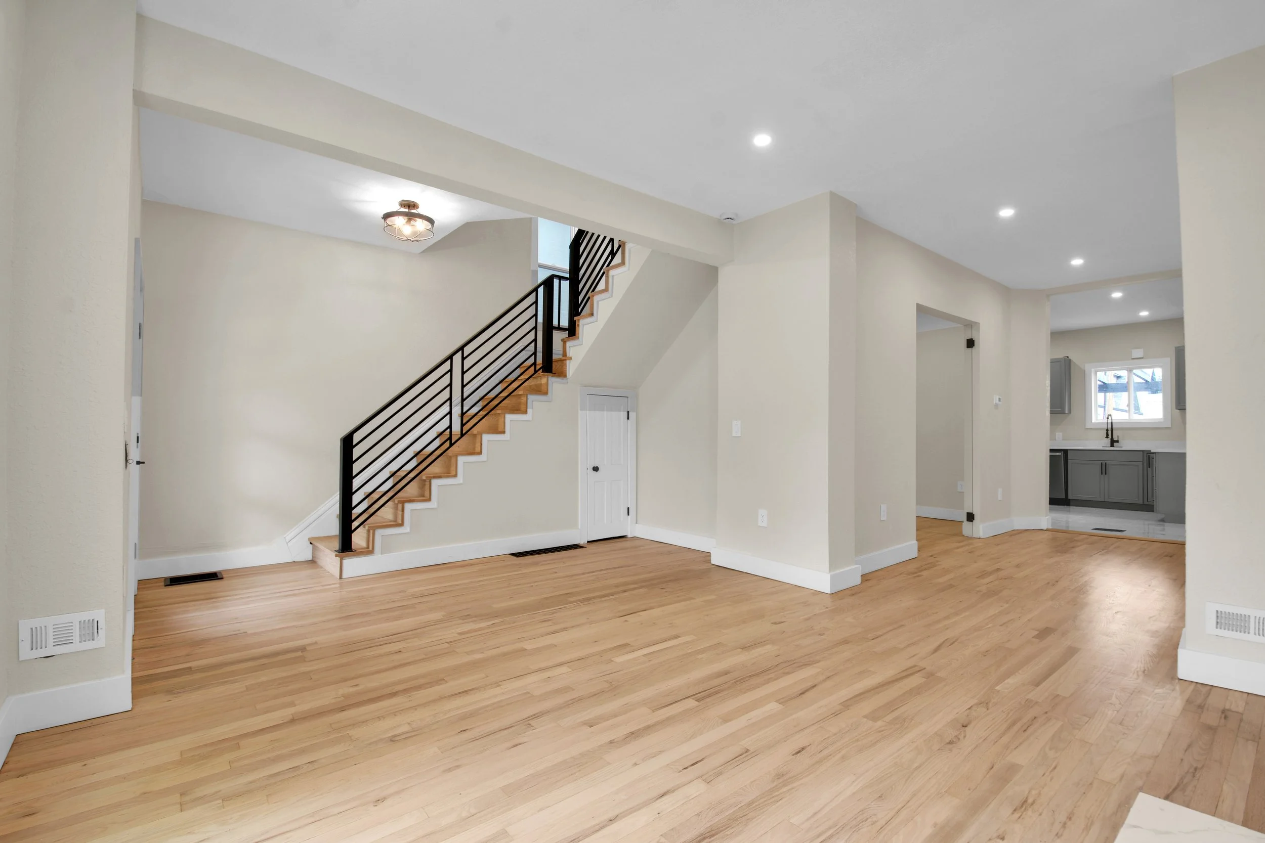 Empty living room with hardwood floors, beige walls, staircase with black metal railing, and small kitchen area in the background.