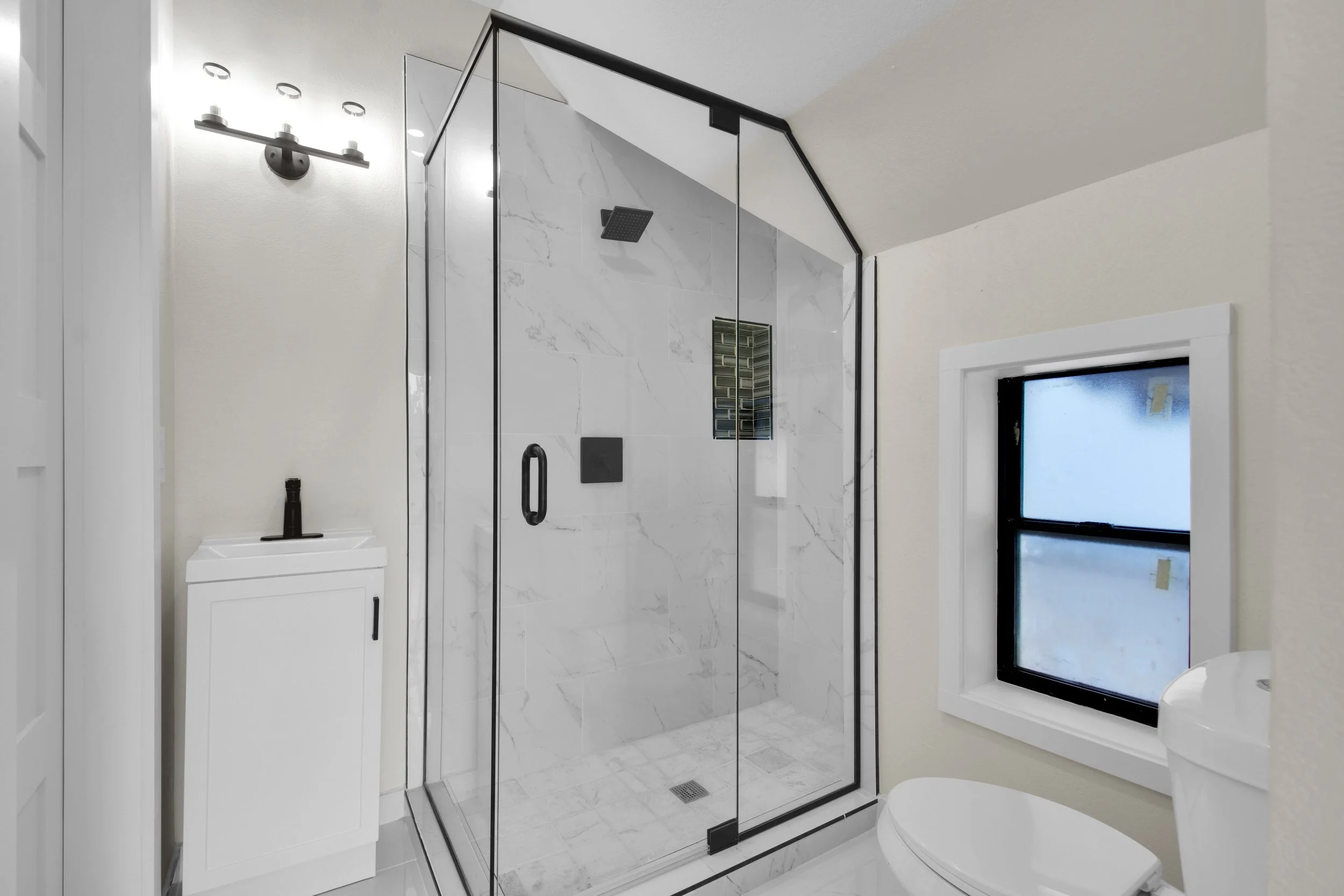 Modern bathroom with a glass-enclosed shower, white walls, a small white vanity, and a frosted window.