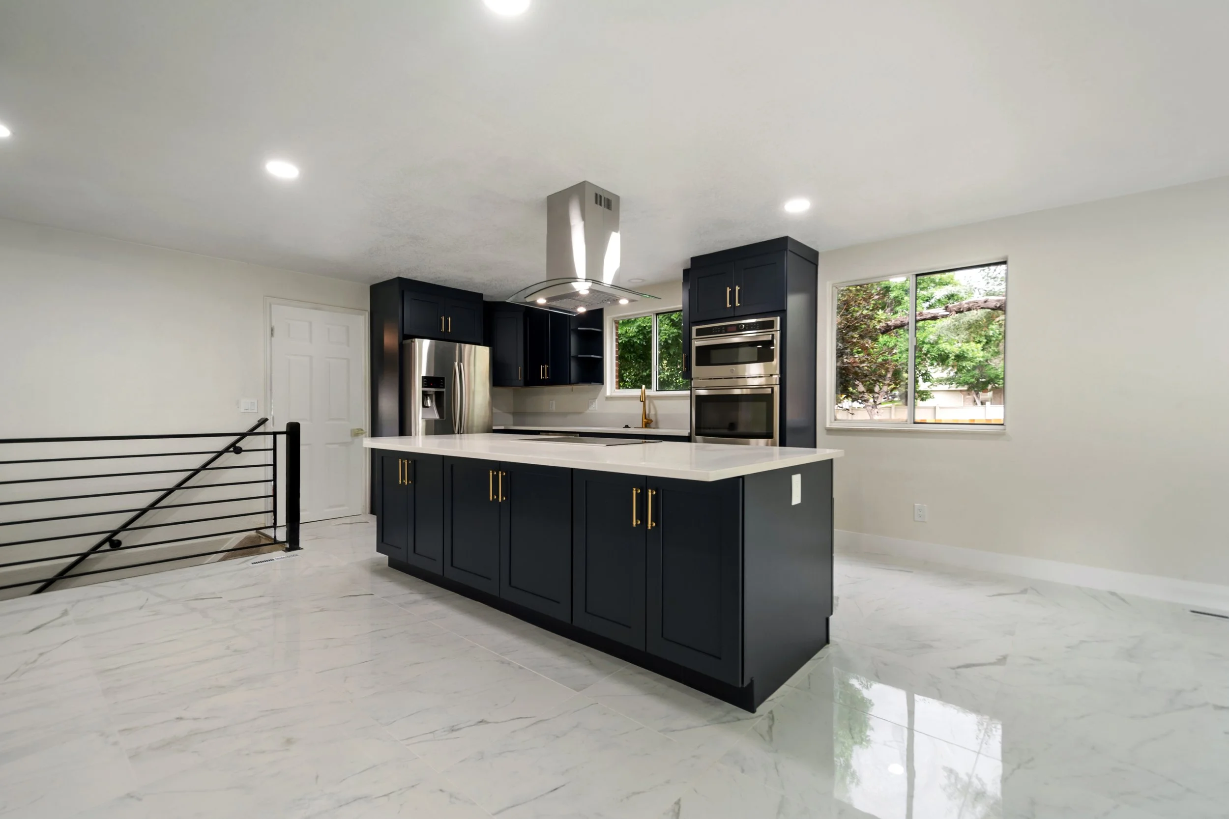 Modern kitchen with dark cabinets, white marble floor, stainless steel appliances, and large windows letting in natural light.