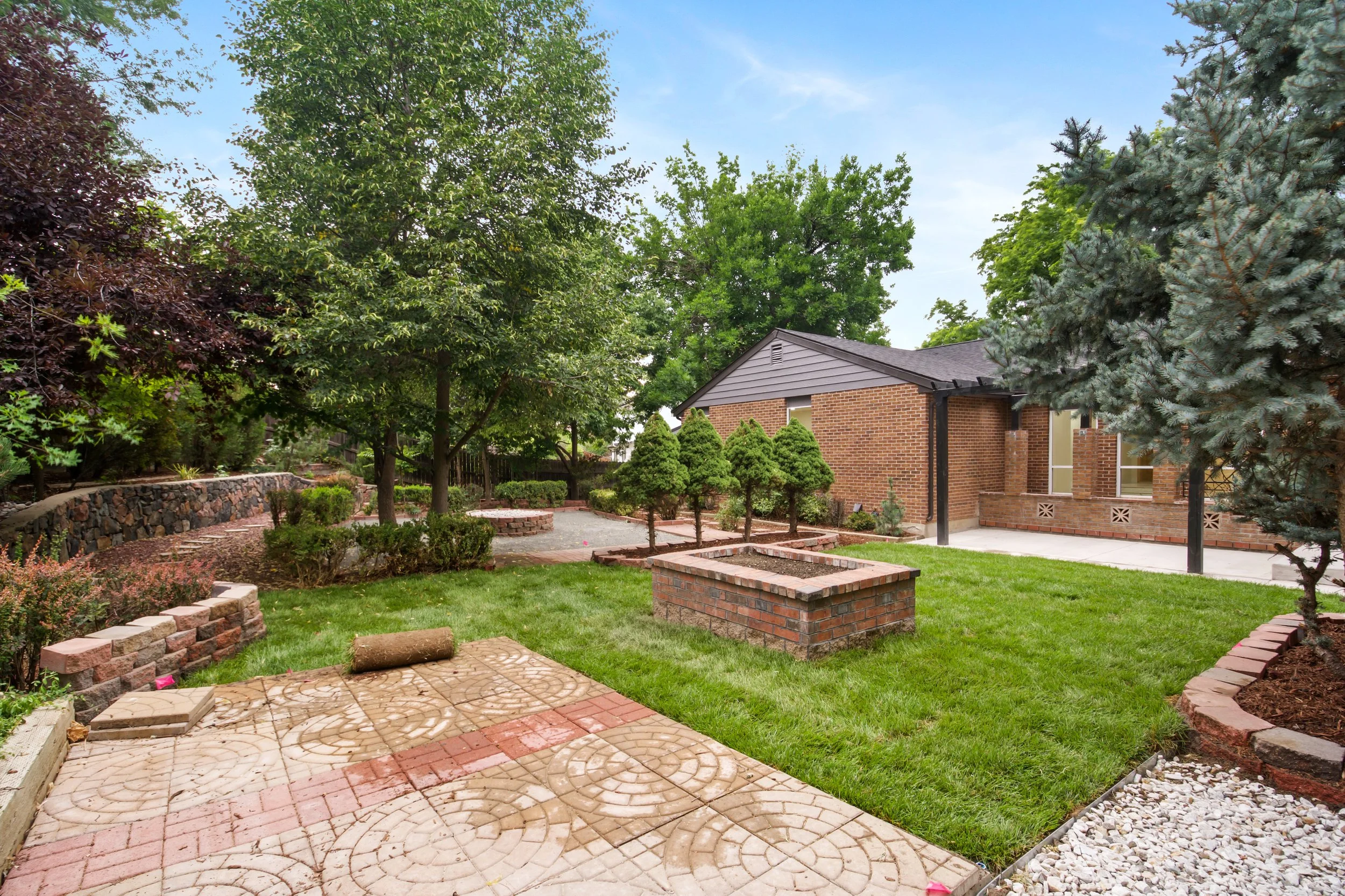 A backyard with green grass, trees, shrubs, brick planters, a stone patio, and a brick house with a gray roof.