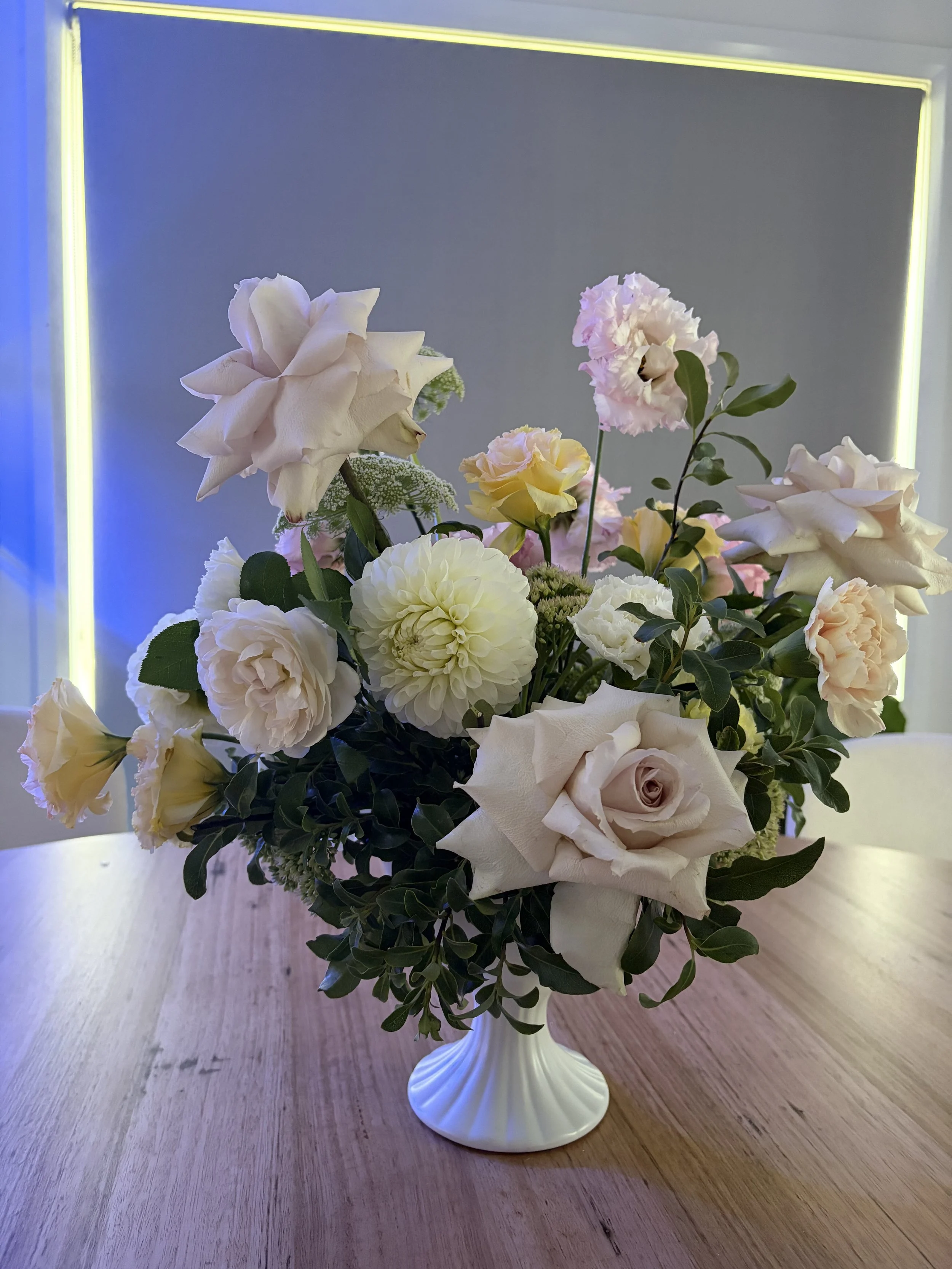 A bouquet of white and soft pink flowers arranged in a white ceramic vase on a wooden table.
