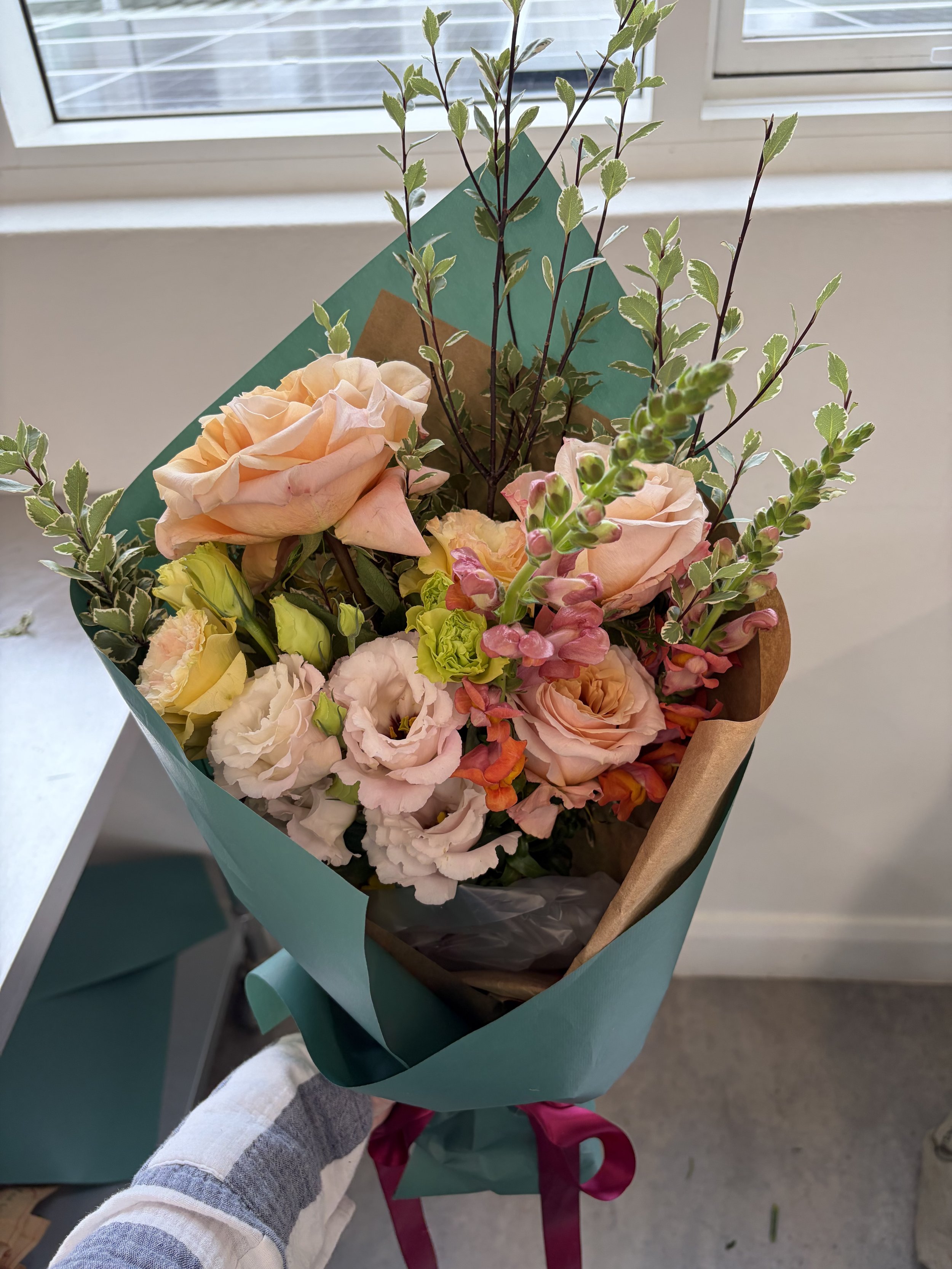 A colorful bouquet of flowers with pink roses, yellow gerbera, and green fillers wrapped in teal paper, held by a person wearing a gray and white striped sleeve, against a background of a window with blinds.