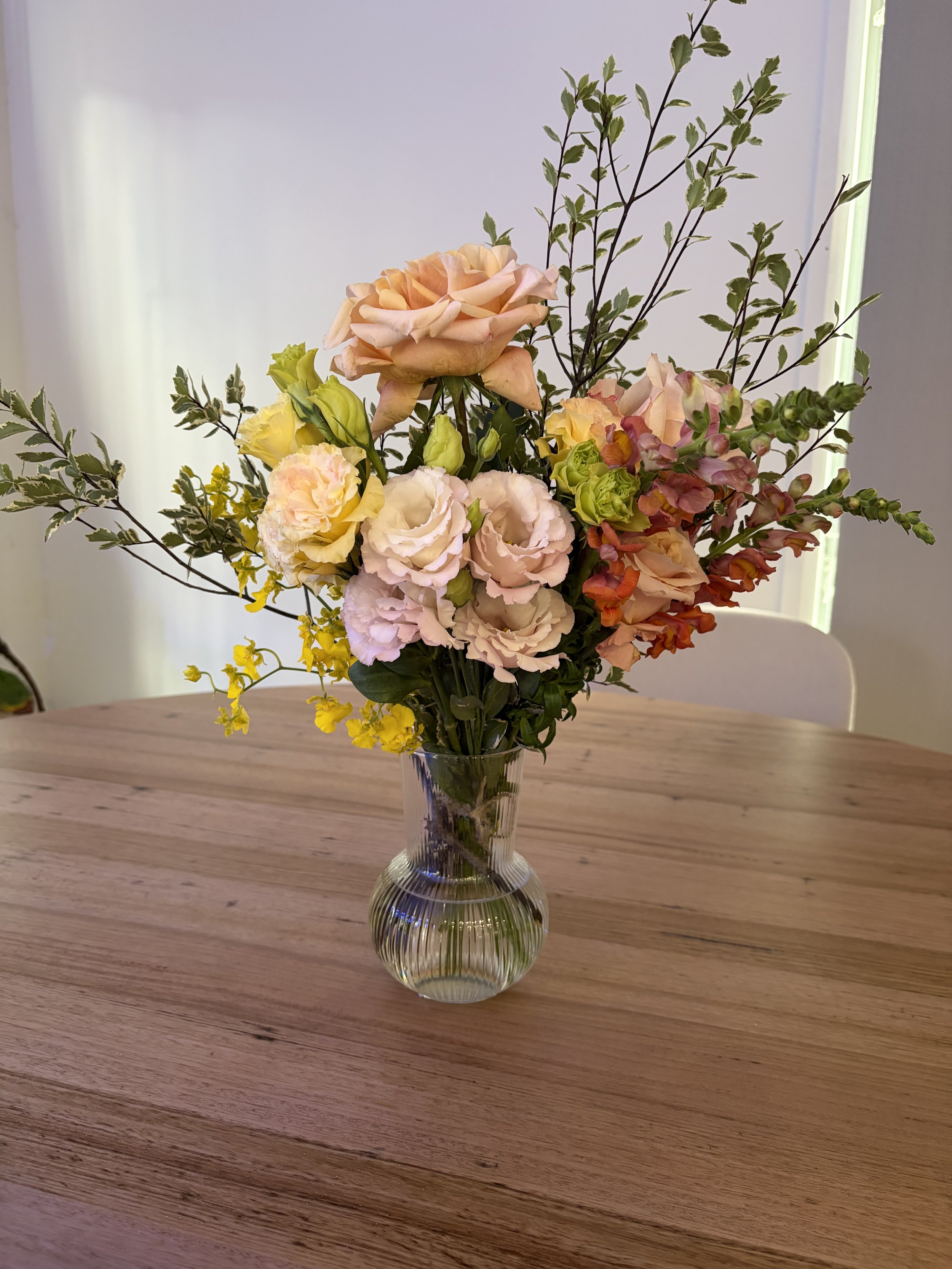 A glass vase with a bouquet of pink, yellow, and peach roses, complemented by green foliage, placed on a wooden table near a window.