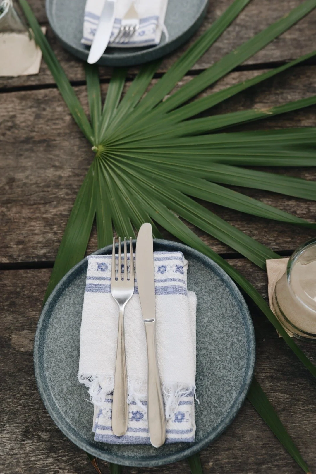 A table setting with a blue plate, silver fork and knife on a folded white and blue striped napkin, a large green palm leaf, a glass of beverage, and a small dish with utensils on a wooden surface.