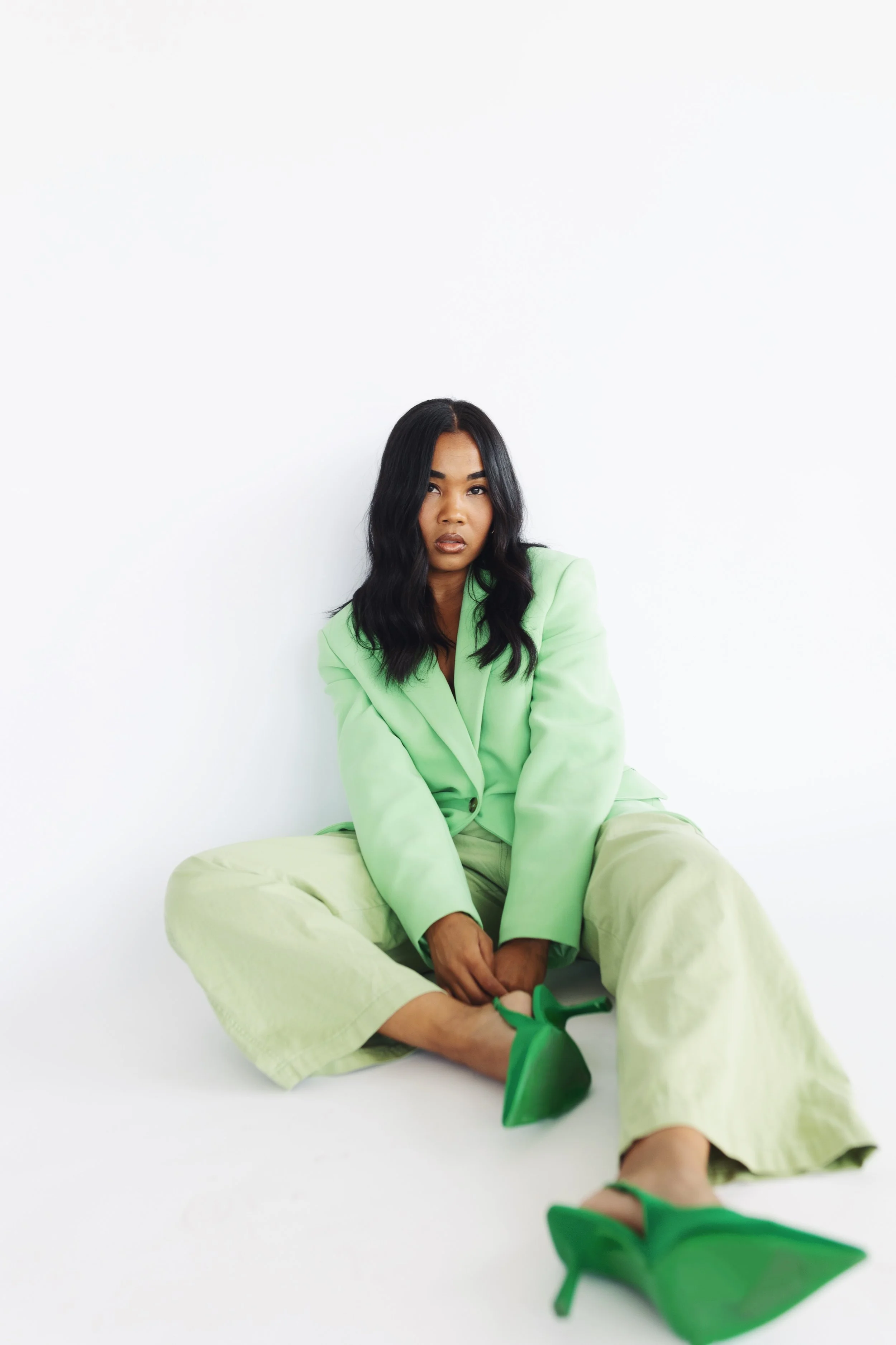 A woman seated on the floor against a plain white background, wearing a light green oversized blazer, green wide-leg pants, and green high-heeled shoes, with dark wavy hair and neutral makeup.
