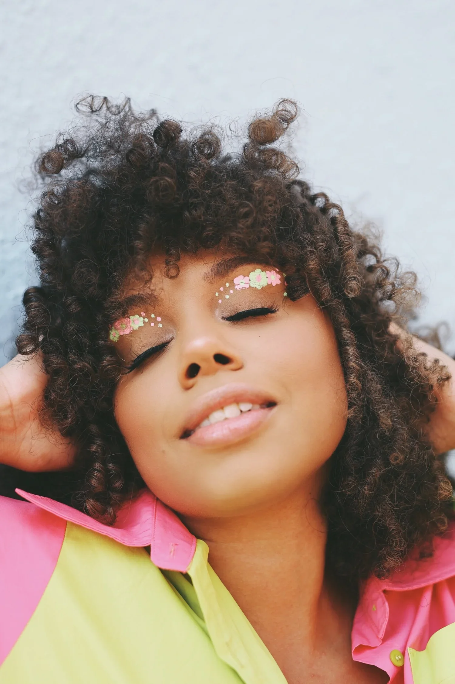 A young girl with curly hair smiling with her eyes closed, wearing eye makeup with floral designs and pink lipstick, dressed in a pink and yellow shirt.