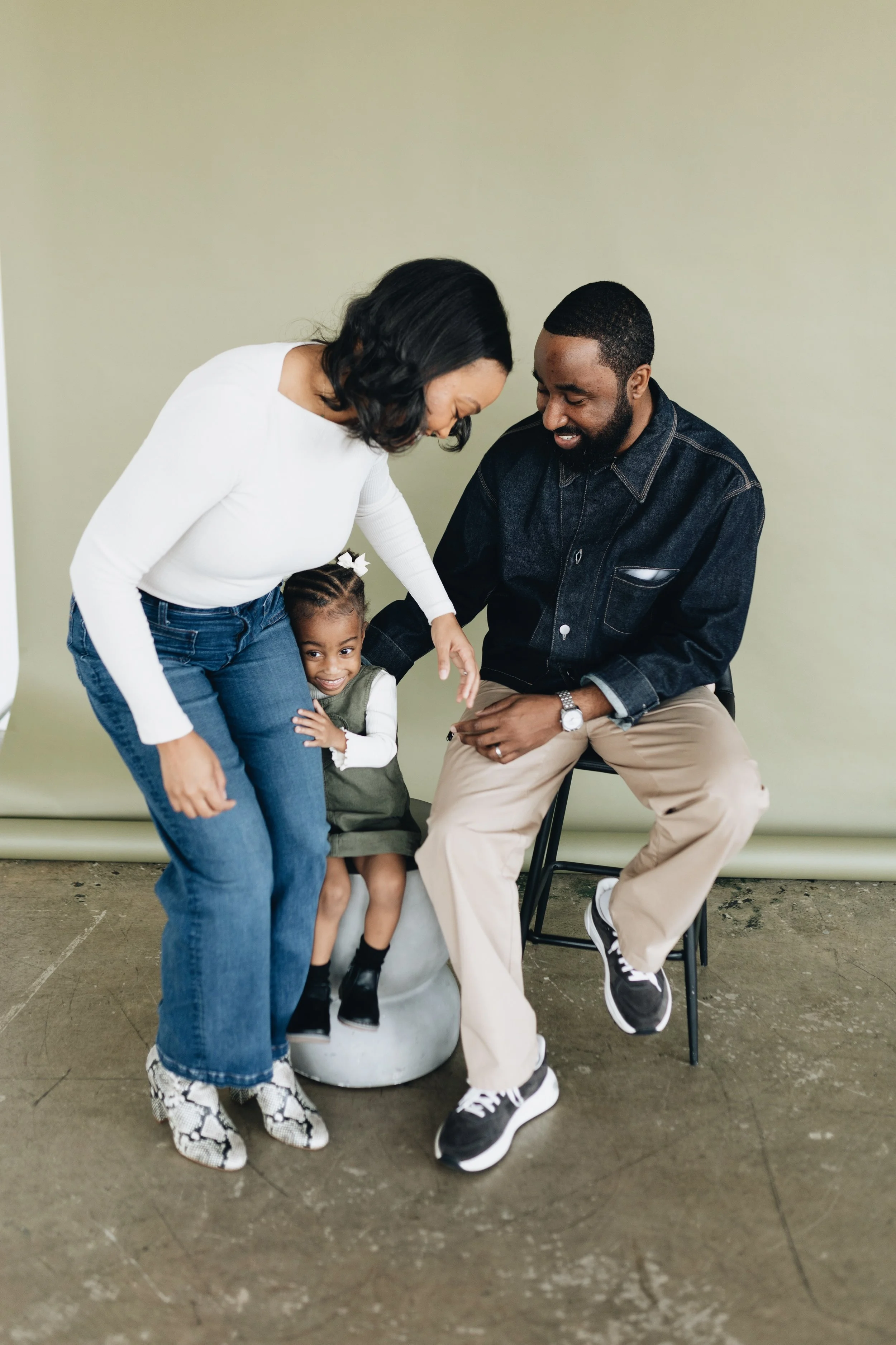 Family of three enjoying a joyful moment together, with a woman and man, and a young girl sitting on a small stool, all smiling and sharing a lighthearted moment.