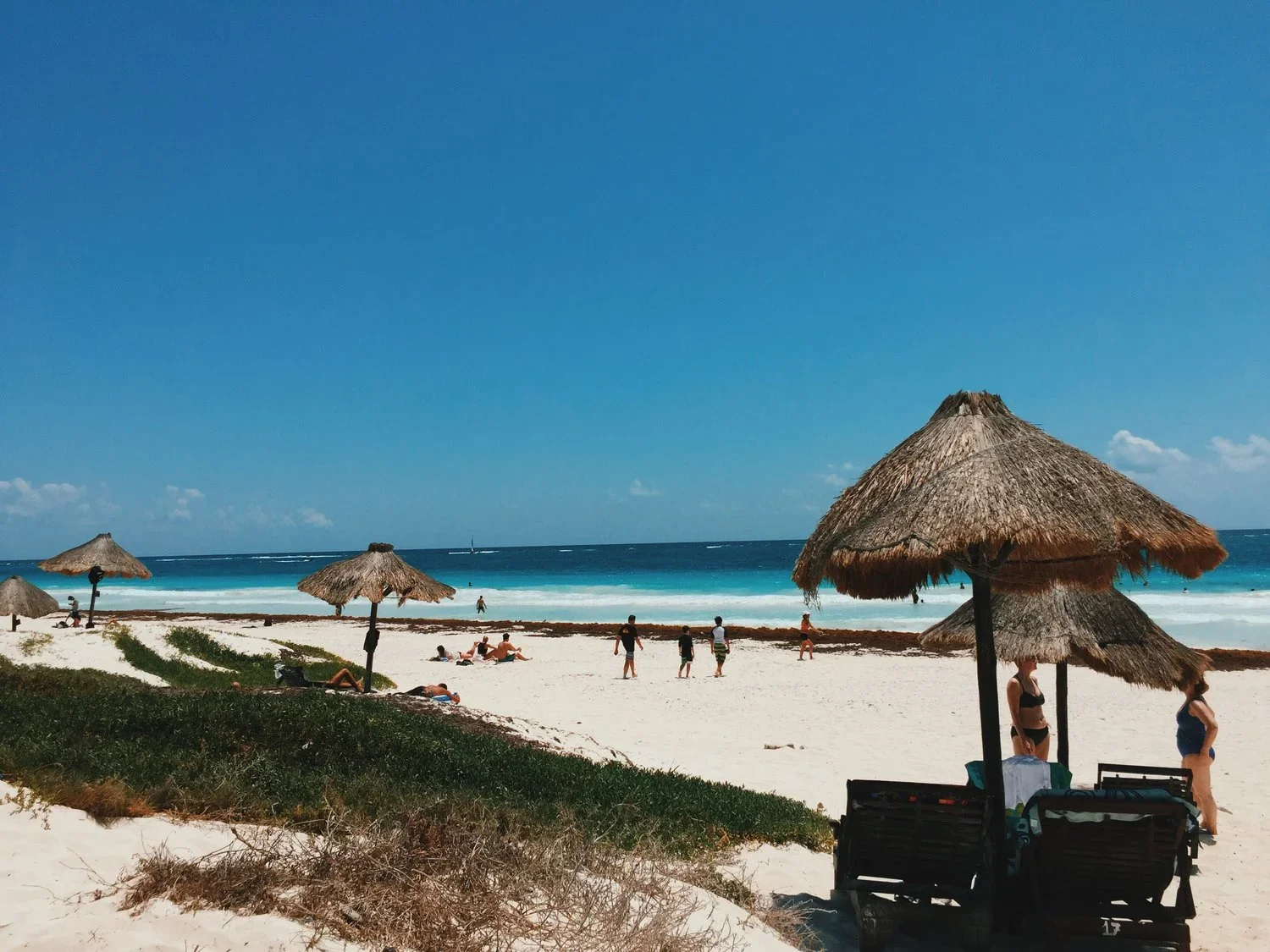 Beach scene with thatched umbrellas, people relaxing on lounge chairs and walking along the sandy shore, and the ocean with blue waves and a clear sky in the background.