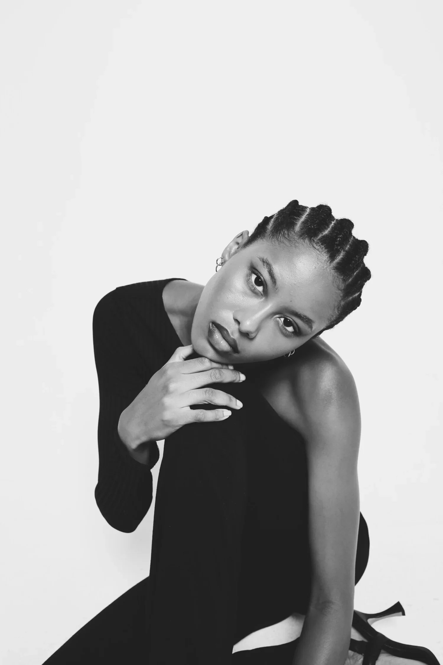 Black and white photo of a woman with cornrow braids, looking directly at the camera with her head tilted, resting her hand on her chin, wearing a dark long-sleeved top, against a plain light background.