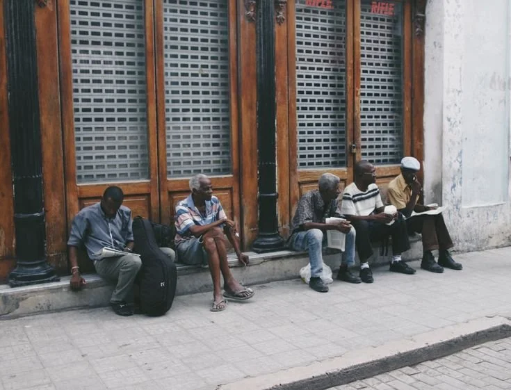 Five men sitting on the sidewalk outside a building with wooden window frames, reading newspapers or looking at papers.