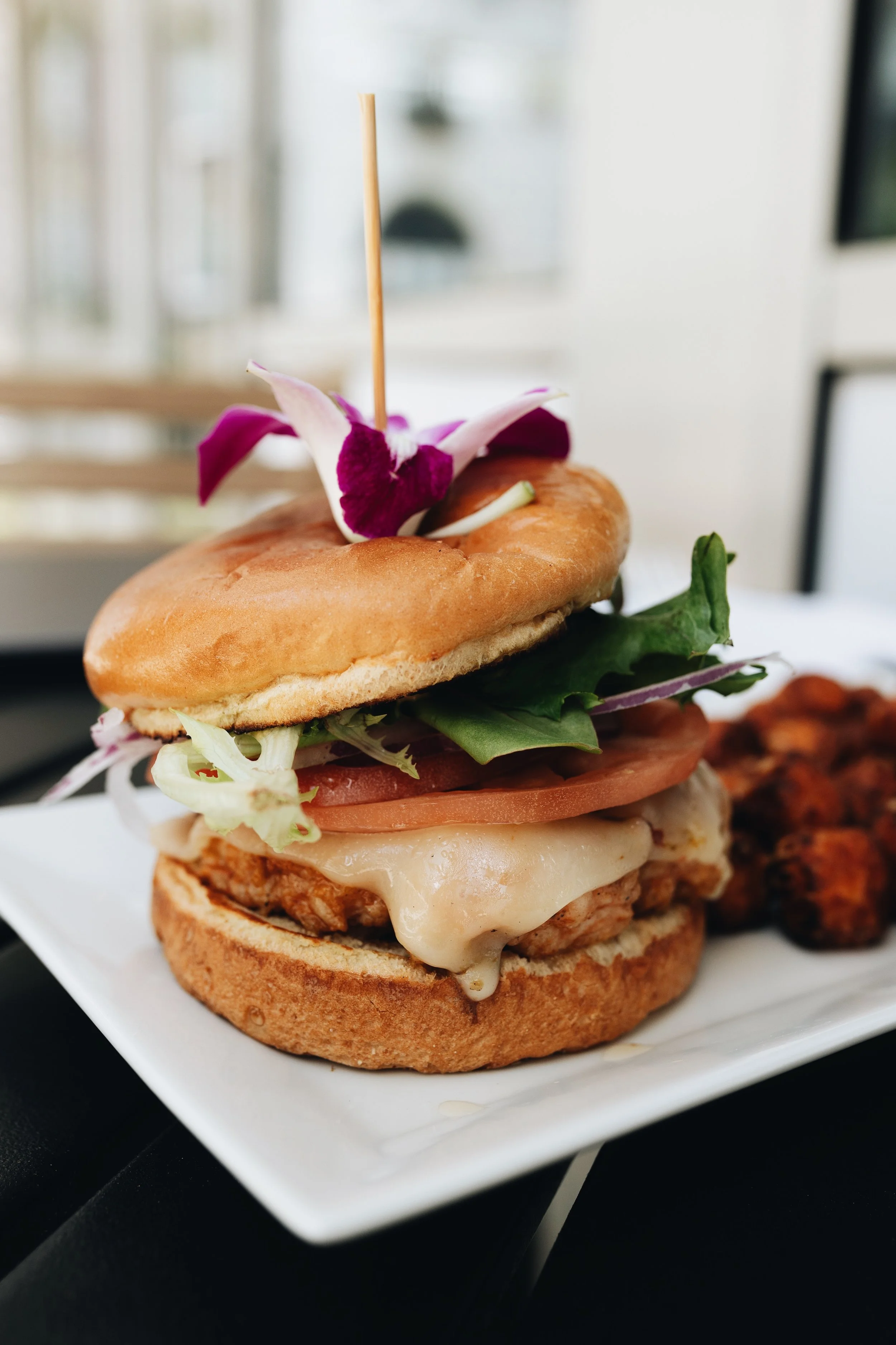 A burger with a bun, cheese, fried chicken patty, tomato, lettuce, and greens, topped with edible flower petals, served with sides of fried food.
