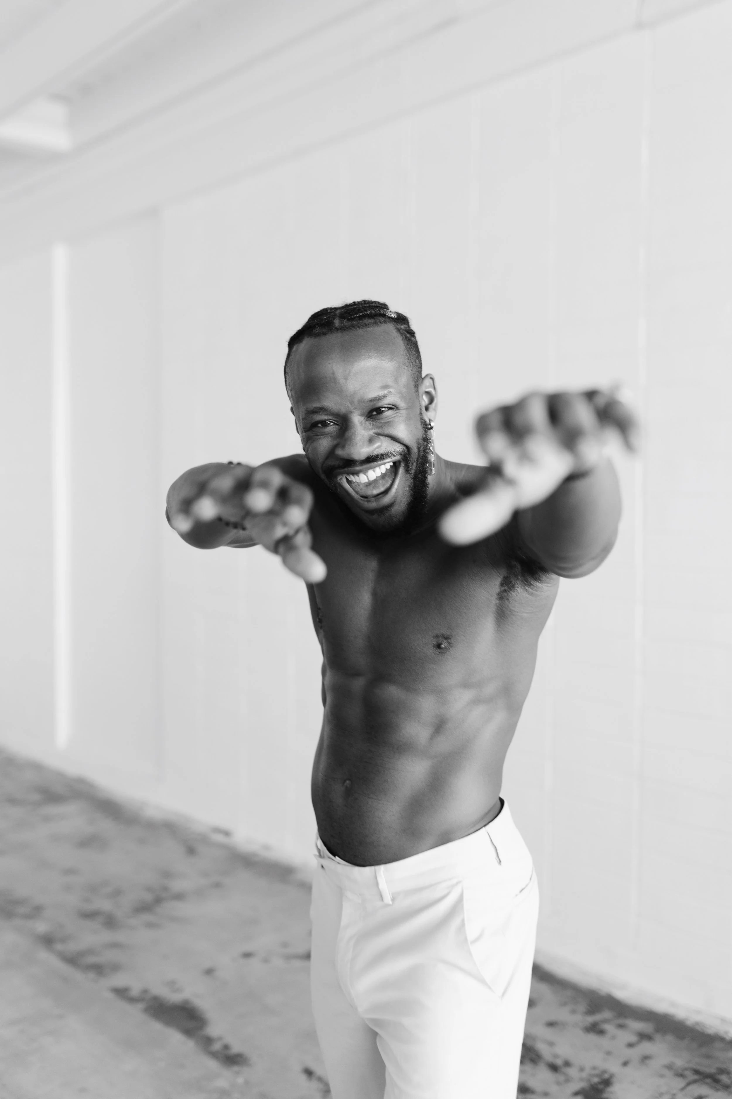 A shirtless man with a beard and braided hair smiling and reaching towards the camera with both hands, standing in front of a plain wall.