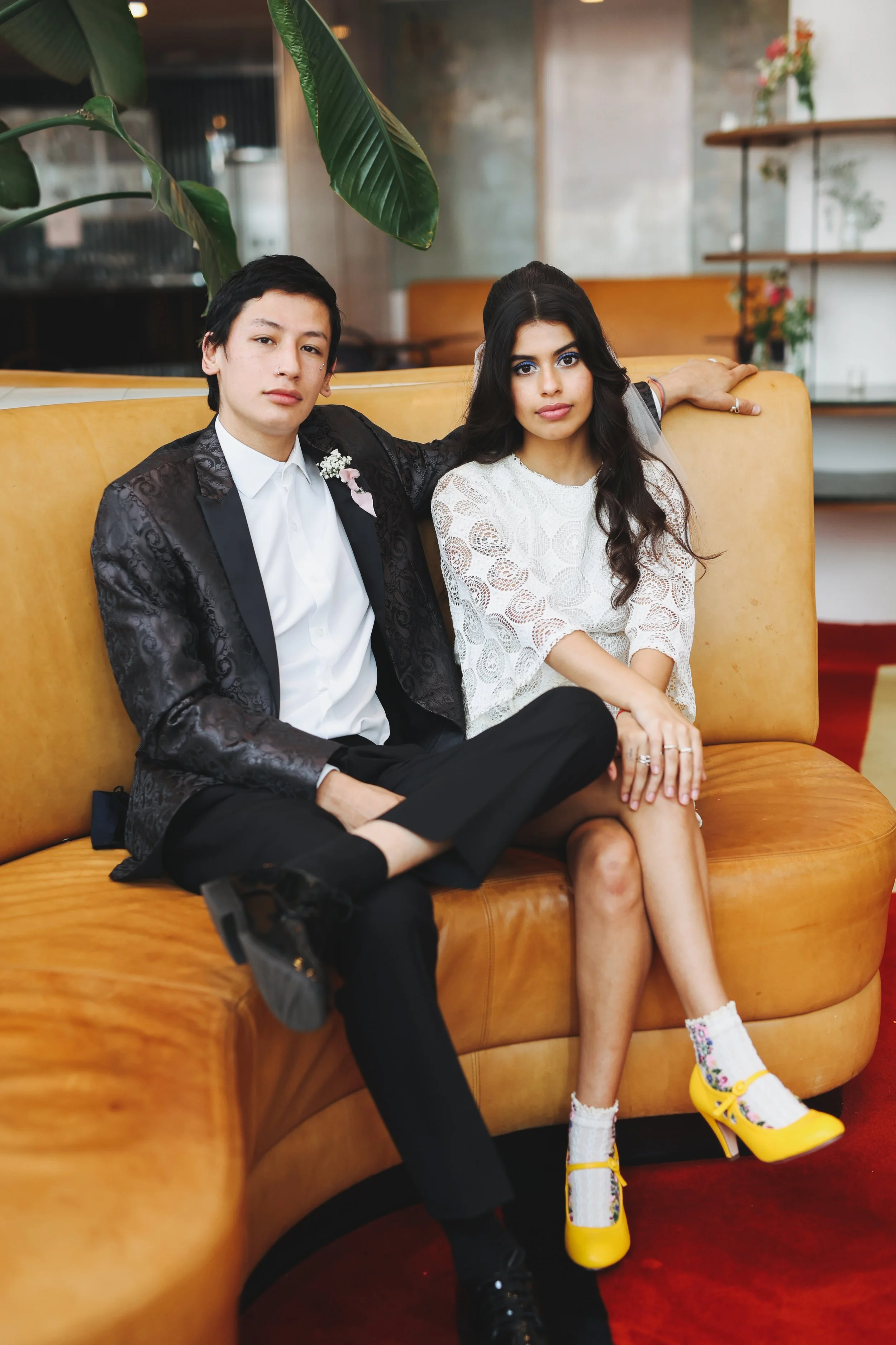 A young man and woman sit on a mustard-colored sofa in a stylish indoor setting, with one arm around each other. The man wears a black patterned suit with a white shirt, while the woman wears a white lace dress with bright yellow high-heeled shoes an