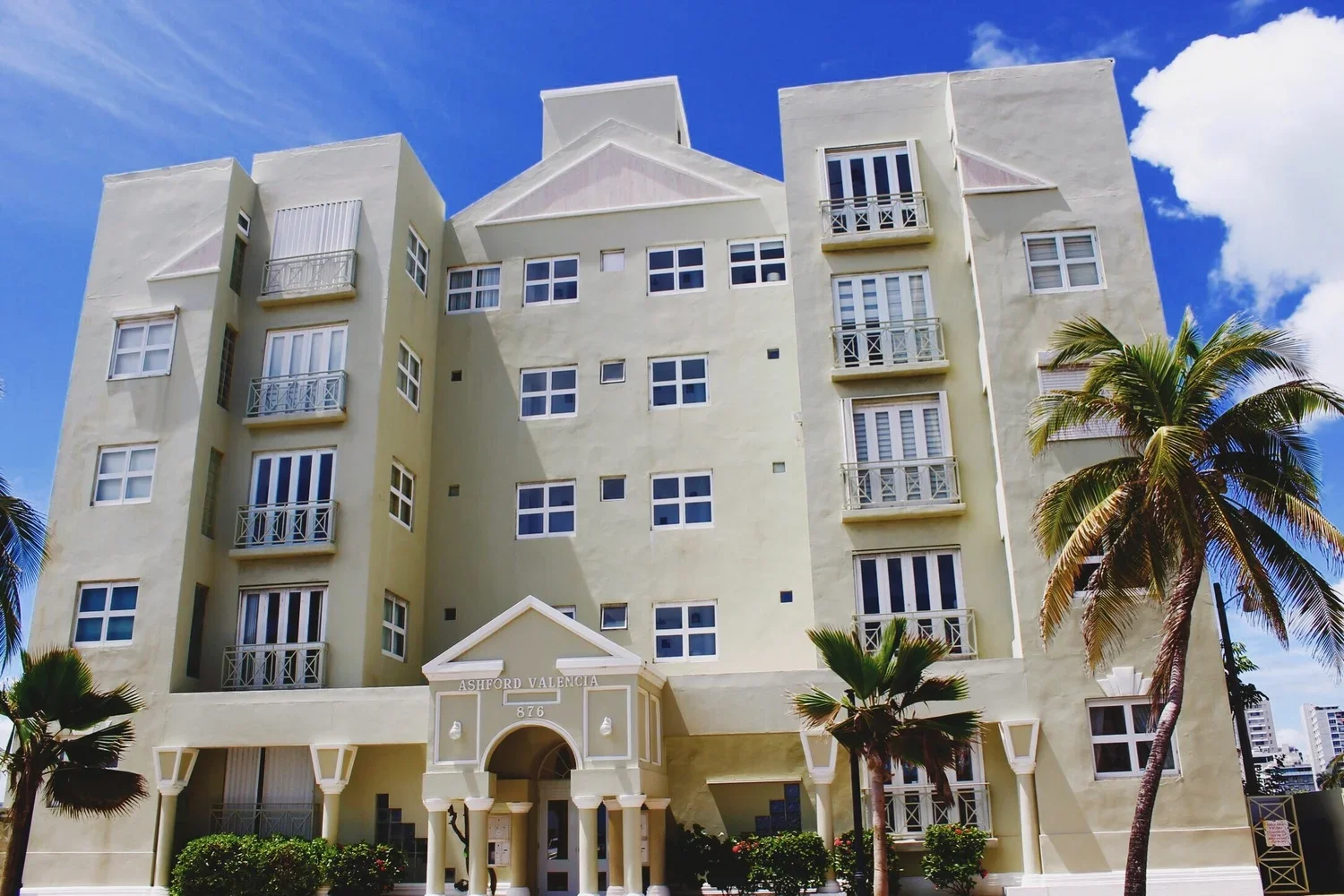 A multi-story apartment building with a light-colored facade, several windows, and small balconies, surrounded by palm trees under a partly cloudy sky.