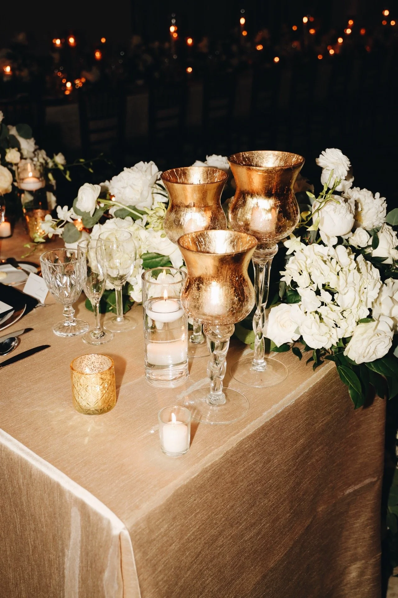 Elegant table decorated with white flowers, gold vases, candles, and glassware at a nighttime event.