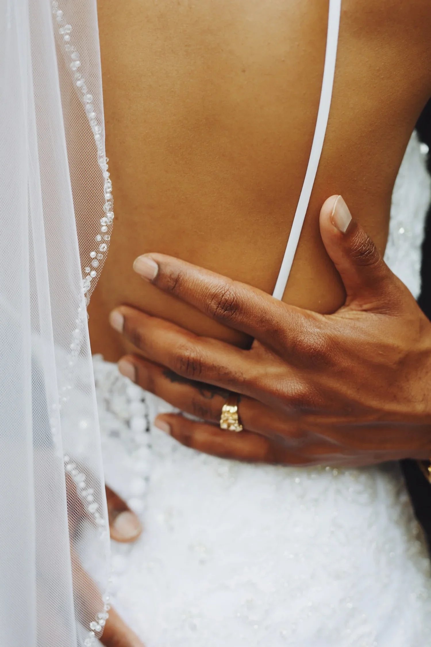 Close-up of a person with darker skin wearing a wedding band on their ring finger, touching their back with their hand, and a portion of a white wedding dress with lace and bead details visible on the shoulder.