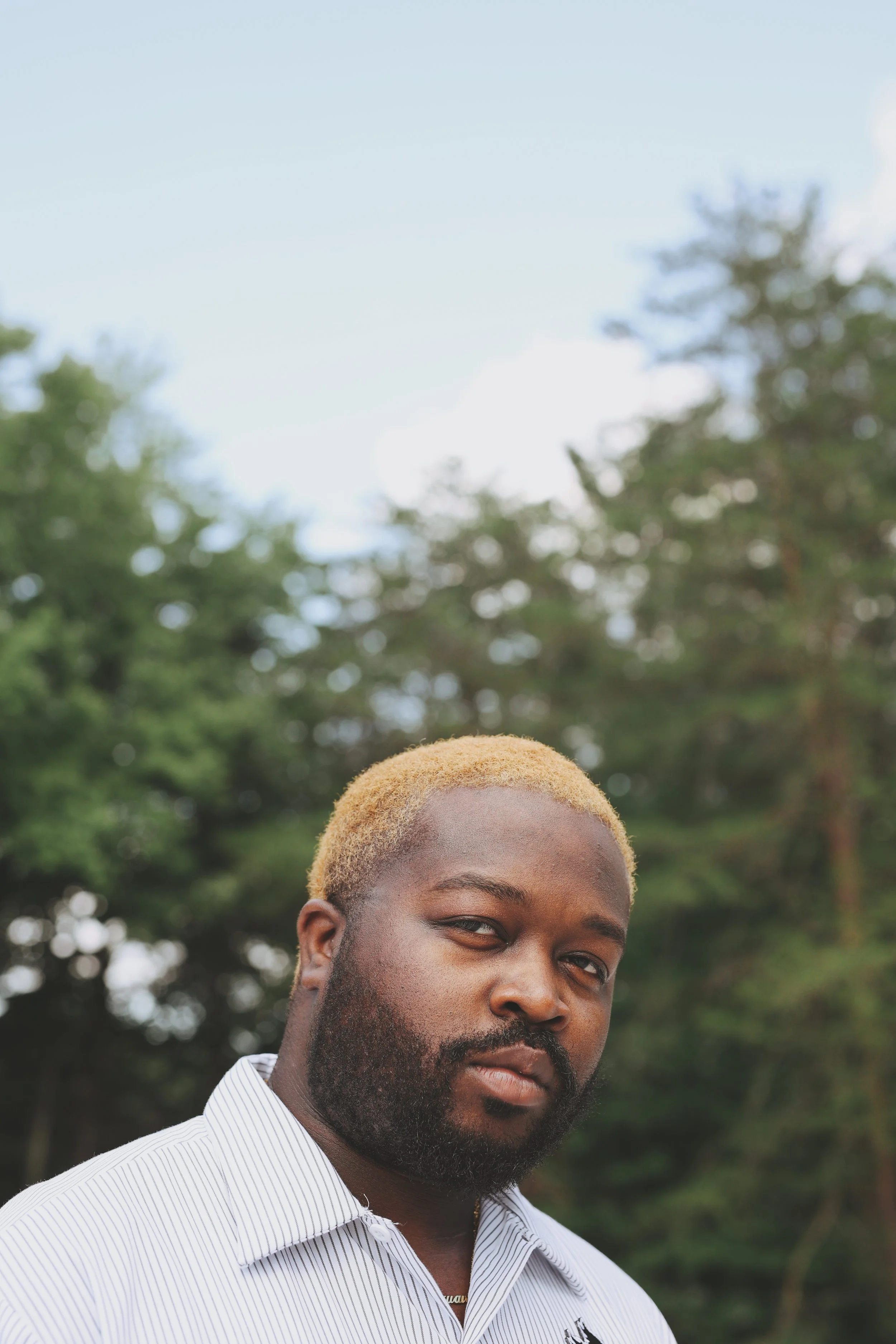 Portrait of a man with blond hair and a beard, wearing a striped shirt, standing outdoors with trees and sky in the background.