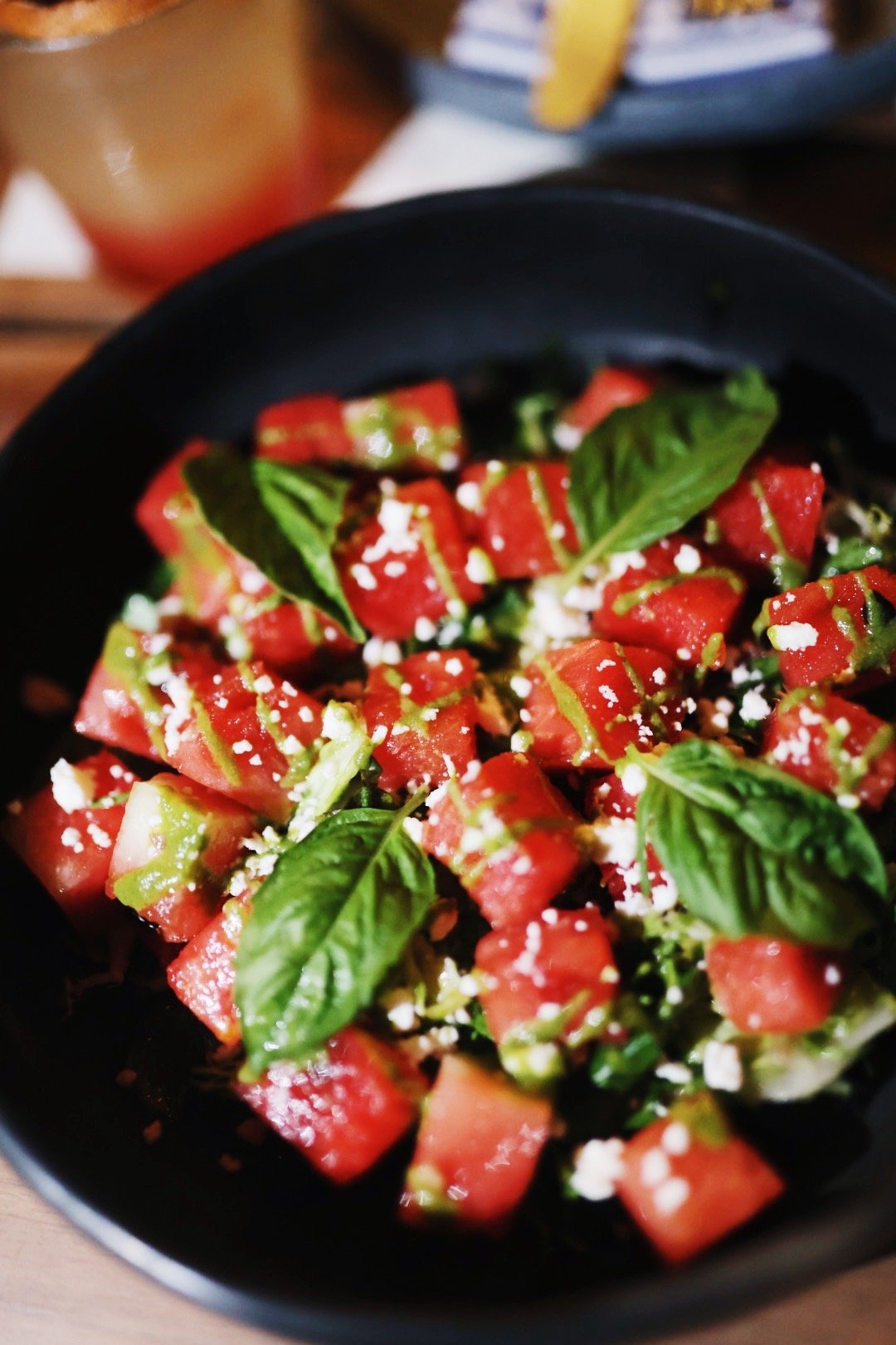 A black bowl of watermelon and tomato salad topped with fresh basil leaves and crumbled feta cheese.