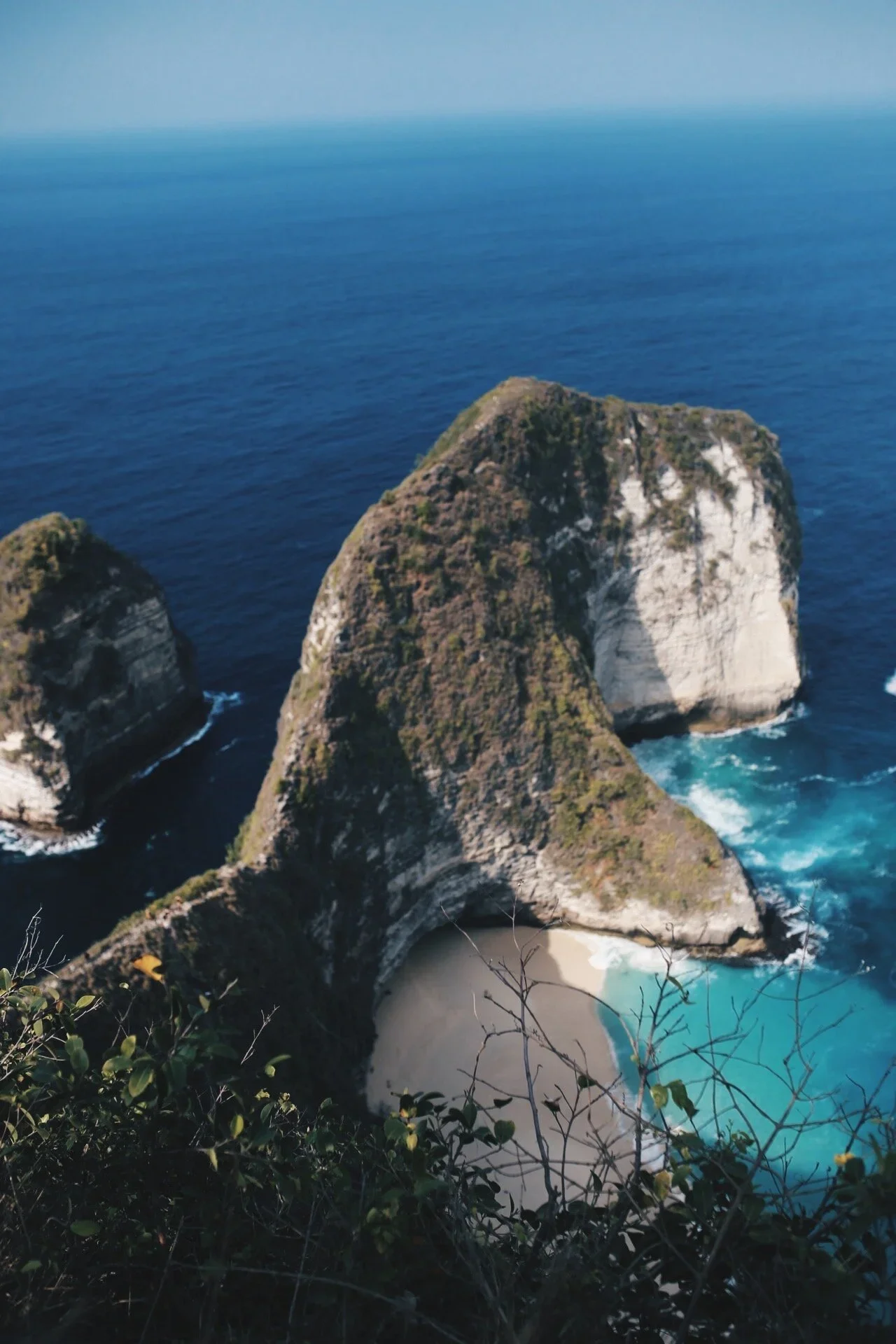 Aerial view of a large, steep rocky island covered in greenery, surrounded by blue ocean water with waves crashing against its base, and a sandy beach in a cove at the island's base.