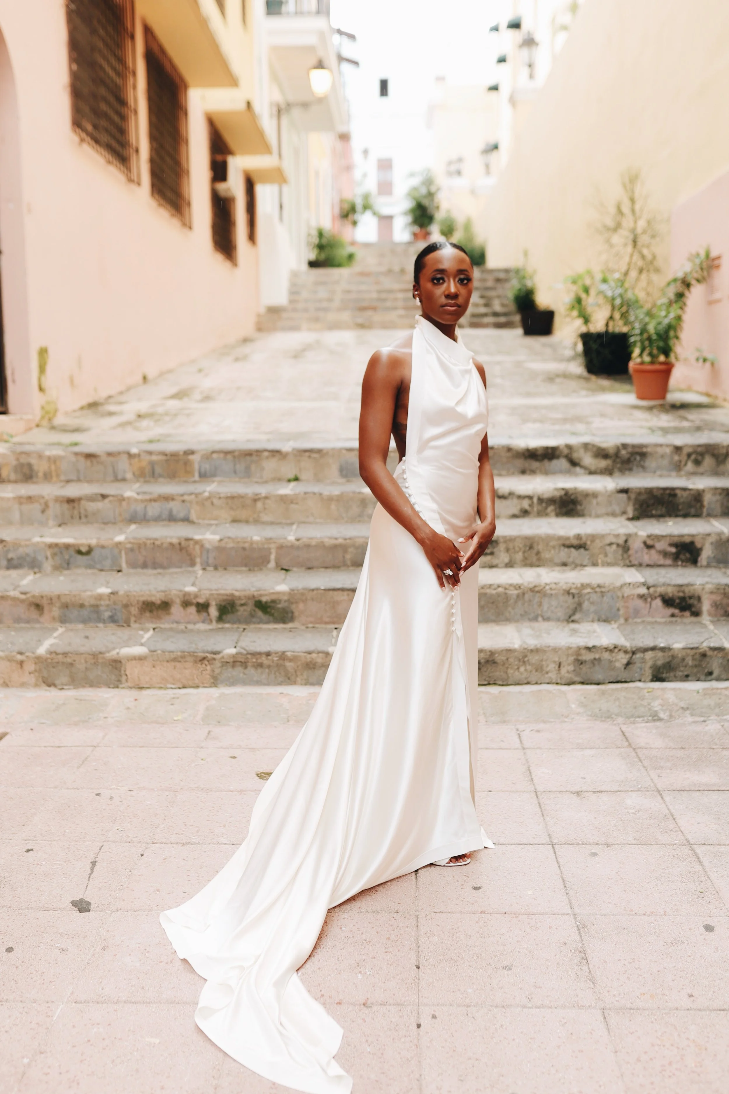 A woman in a white satin gown standing on an urban sidewalk with stairs and potted plants in the background.