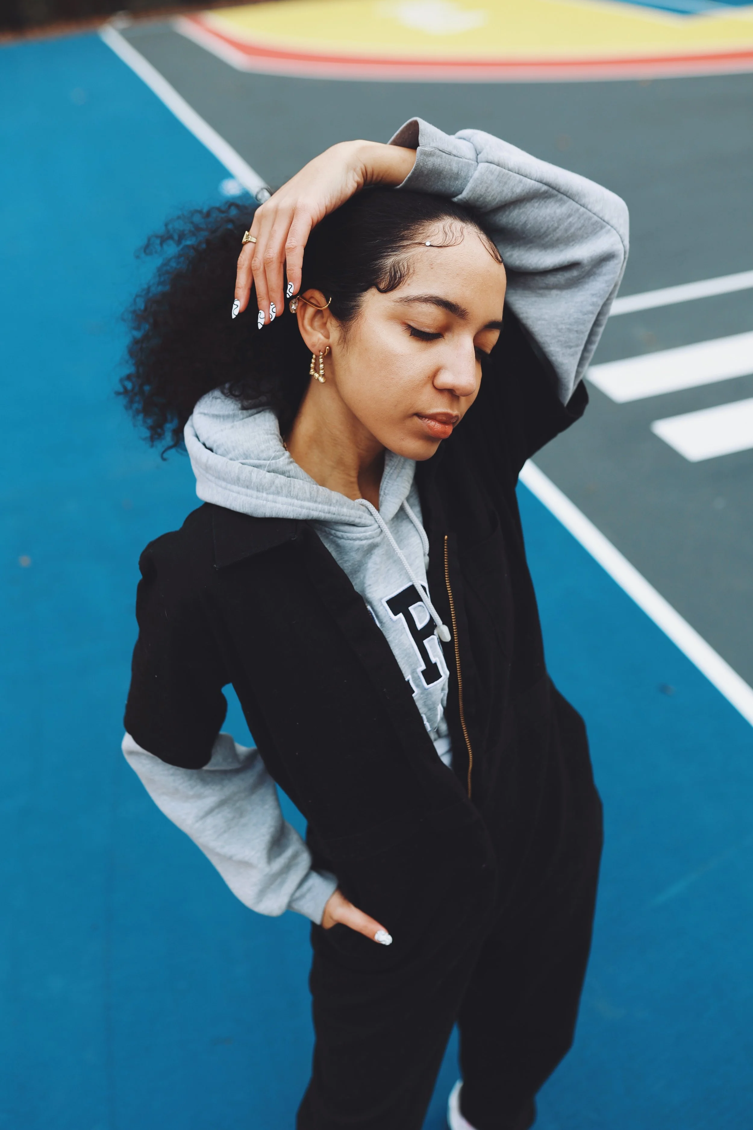 Young woman with curly hair and multiple earrings poses with one hand behind her head and the other on her hip on a colorful outdoor courtside.