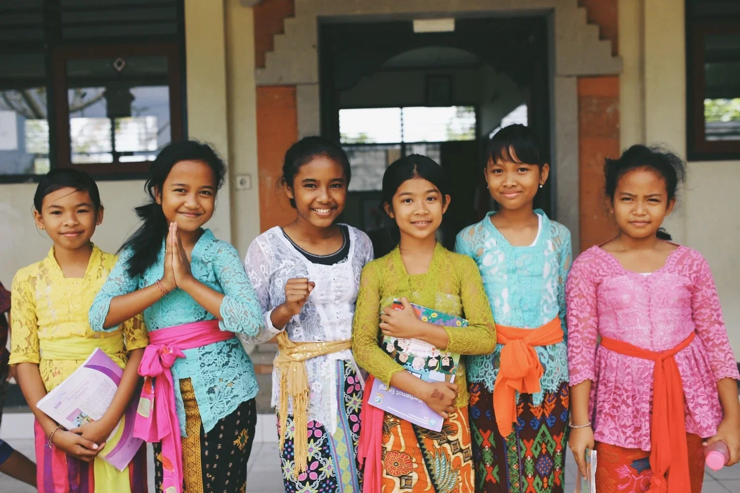 Seven young girls standing together in front of a building, dressed in colorful traditional clothing, holding books and smiling at the camera.