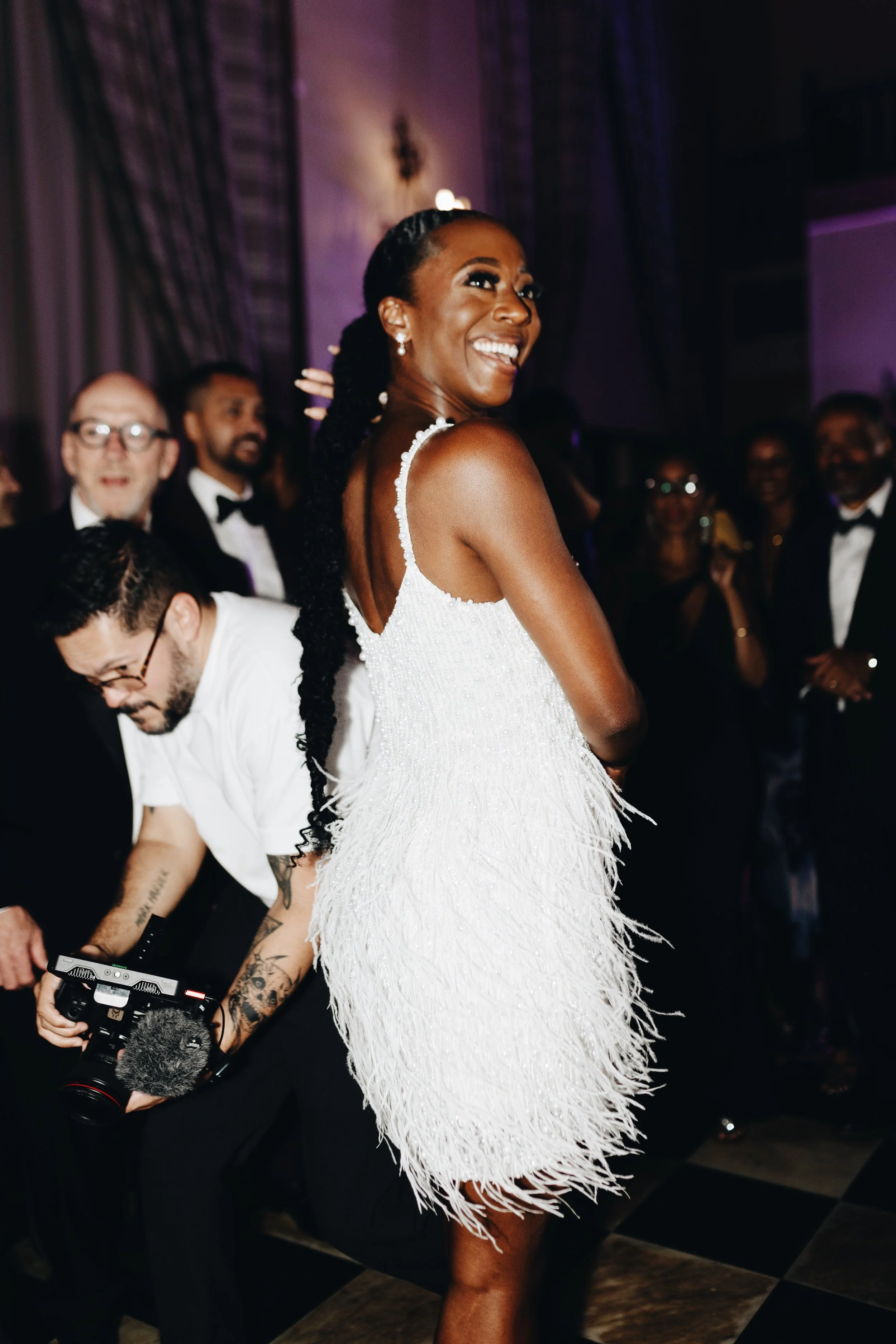 A woman in a white dress with fringe details smiling at a formal event, surrounded by other elegantly dressed people.