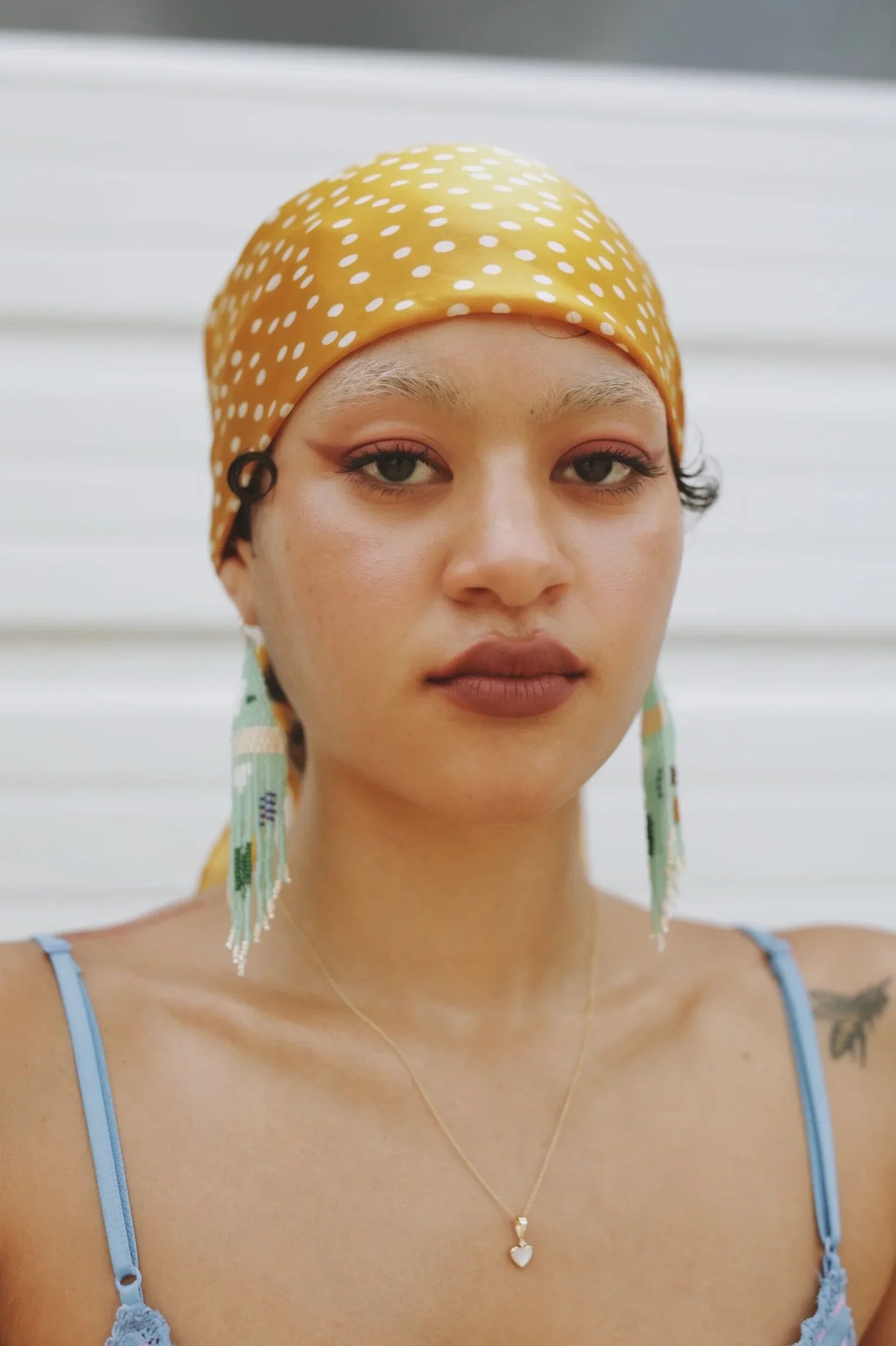 A young woman with makeup and earrings wearing a yellow polka dot headscarf, a blue spaghetti strap top, and a necklace with a heart pendant.