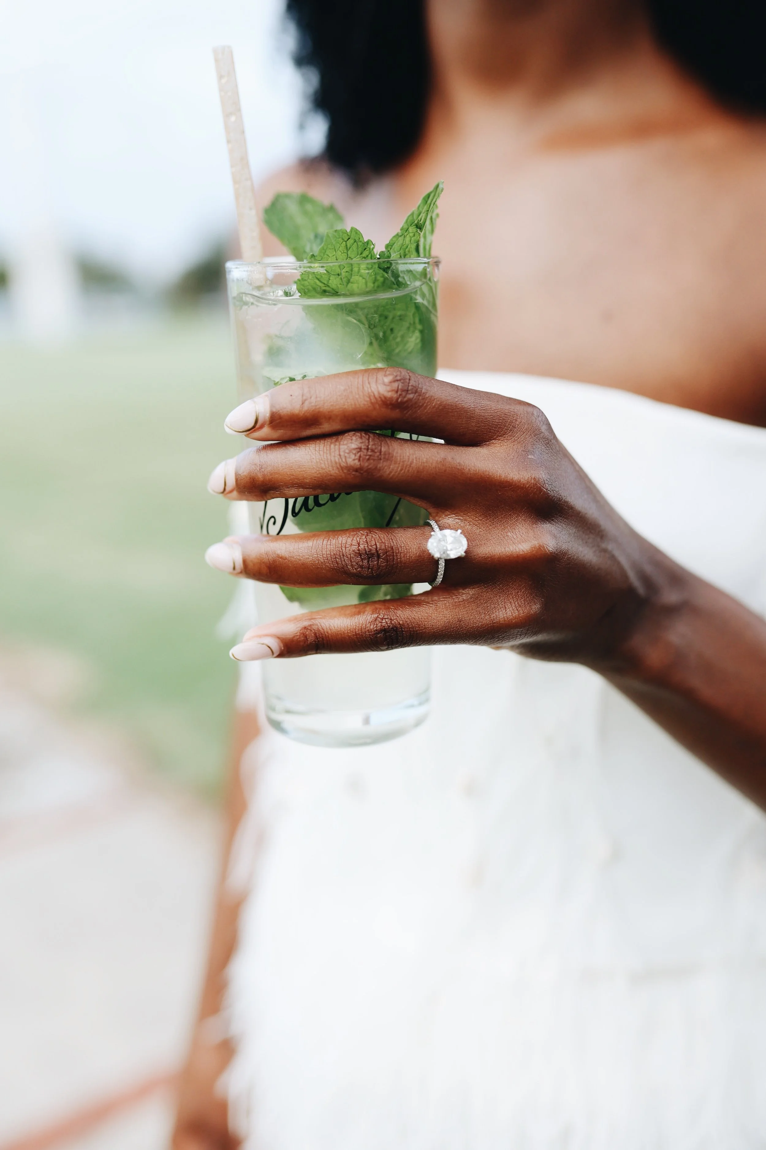 A person with dark skin wearing a white dress holding a glass of mojito cocktail with mint leaves and ice, showing an engagement ring with a large gemstone on their left hand.