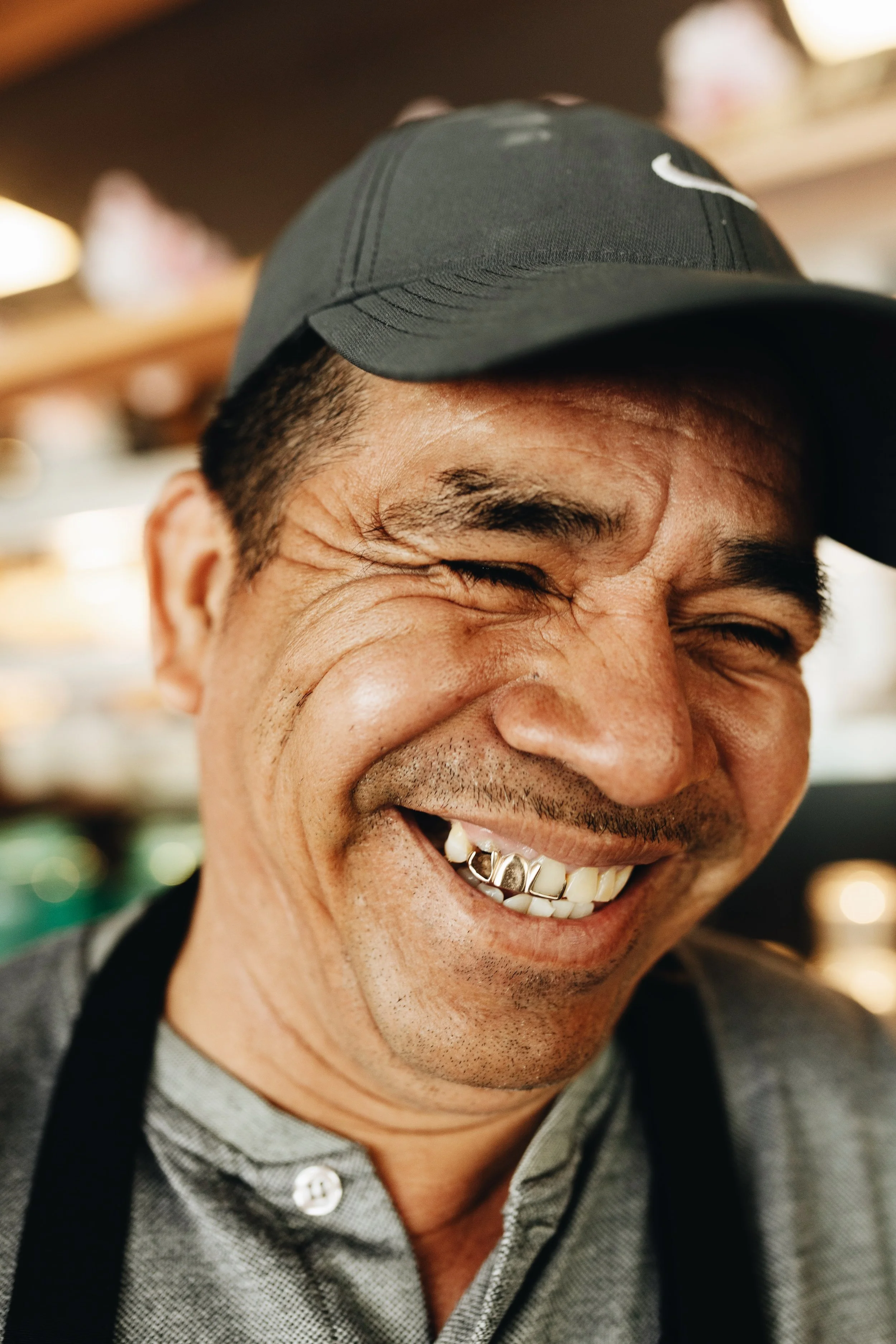 A man smiling widely, showing his teeth with a gold grill, wearing a black Nike cap.