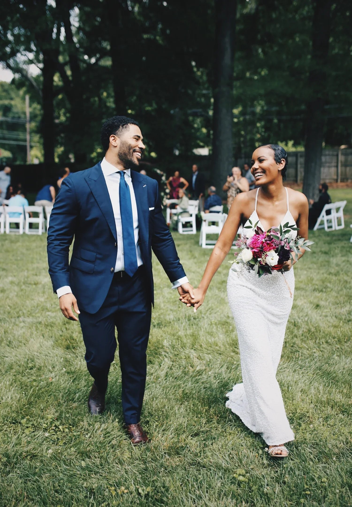 A newlywed couple holding hands and smiling as they walk on grass outdoors at a wedding celebration, with guests seated in the background.