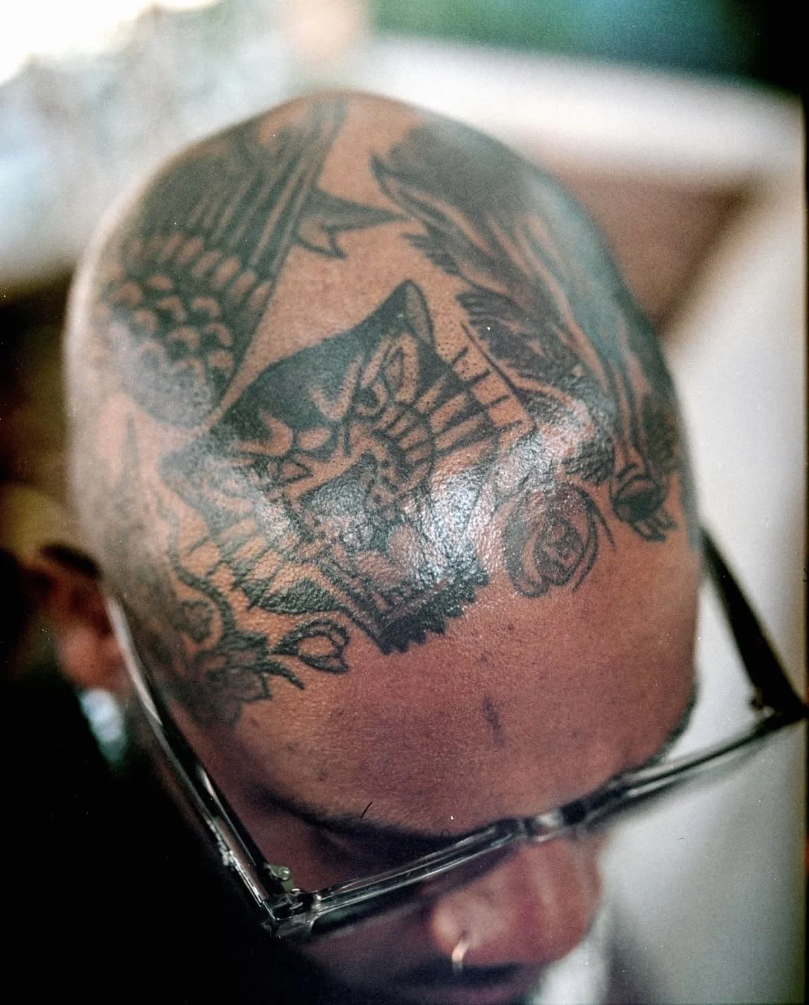 Close-up of a person's bald head with a large tattoo of a cat's face on the crown. The tattoo features a detailed cat face with prominent eyes, nose, and mouth, and surrounding ornamental designs. The person is wearing glasses.