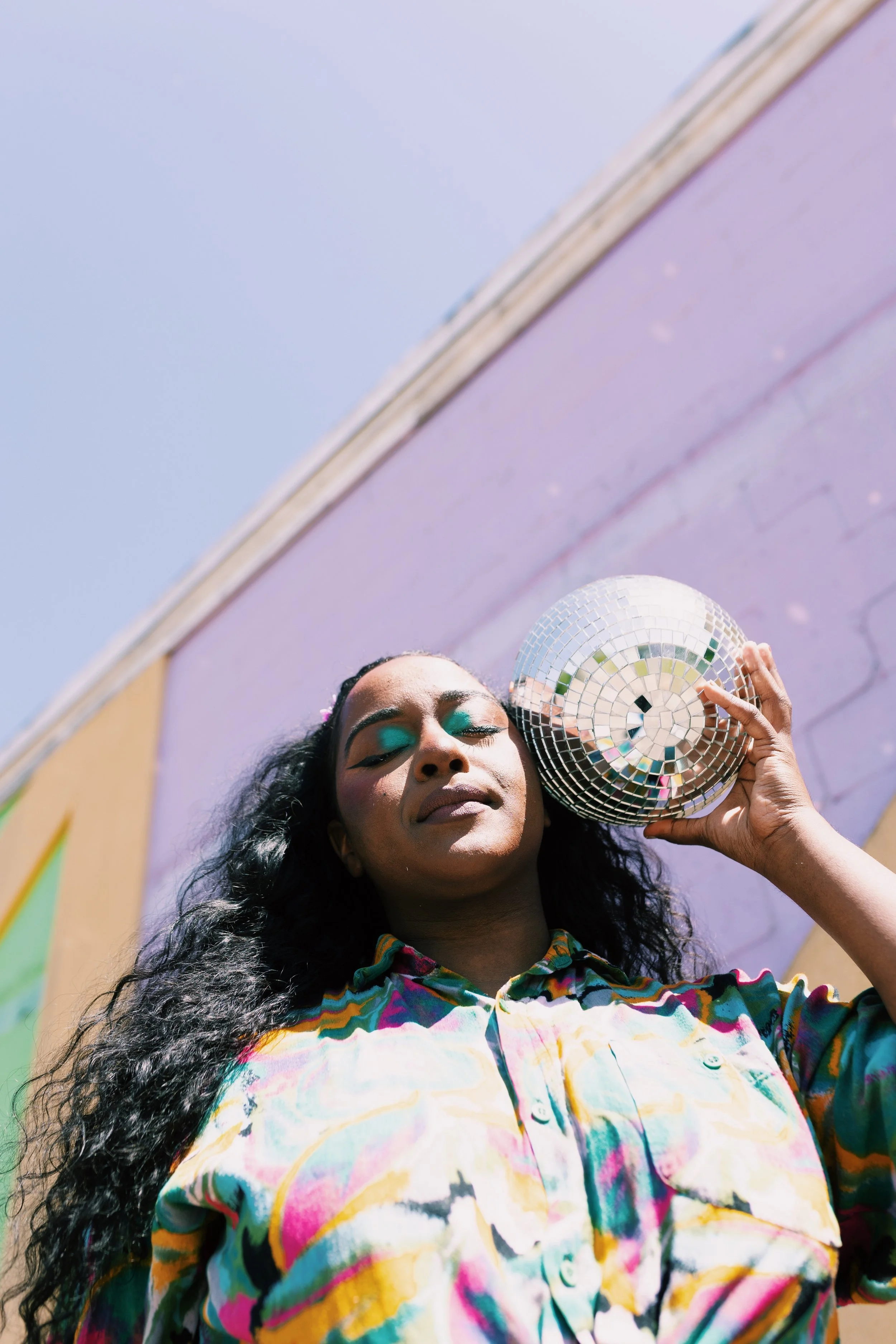 A woman with long curly hair, closed eyes, and colorful makeup, holding a silver disco ball near her ear, standing against a pastel-colored mural on a sunny day.