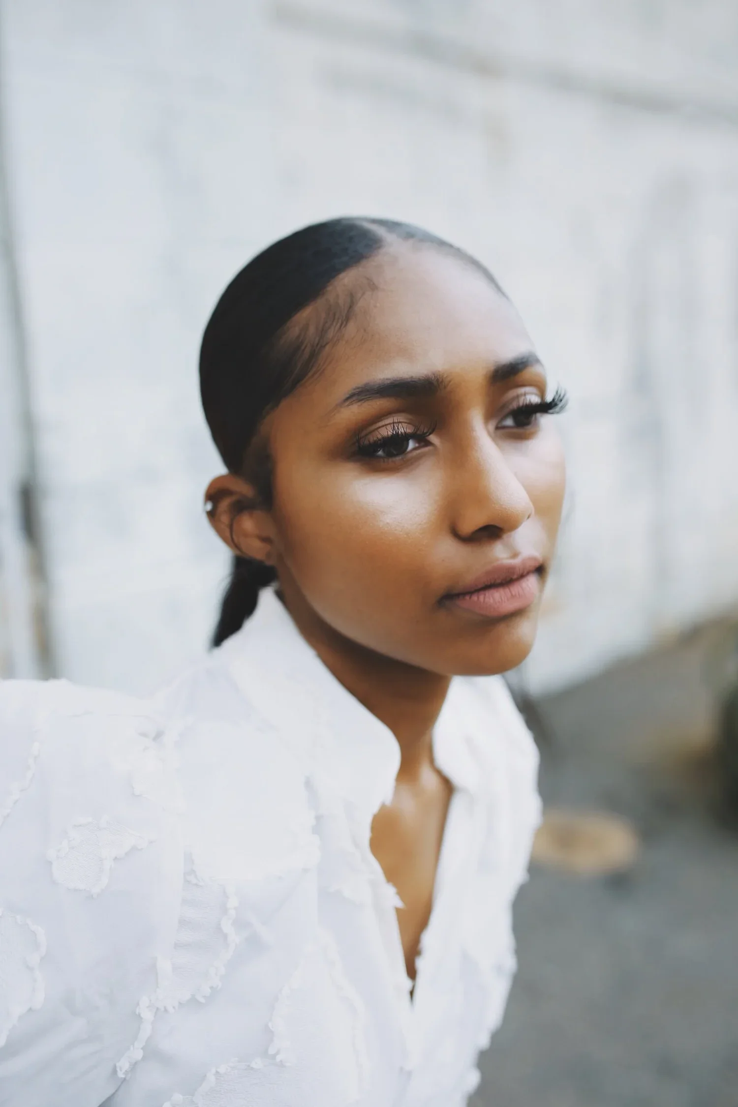 Close-up of a woman with dark hair slicked back, wearing a white textured blouse, standing outdoors against a light-colored wall.
