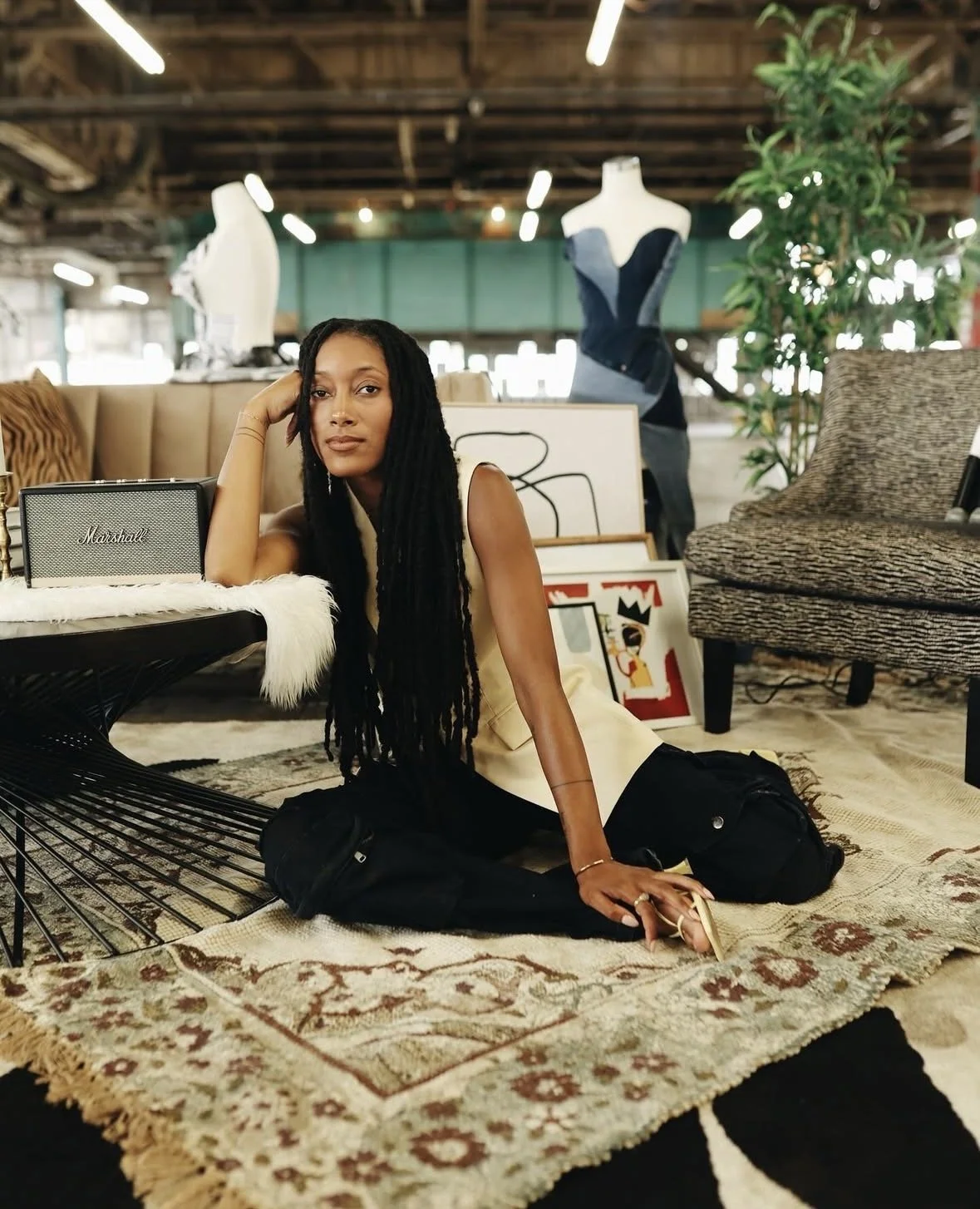 A woman with long black dreadlocks sitting on a patterned rug in an industrial-style space with furniture and art decor around her.