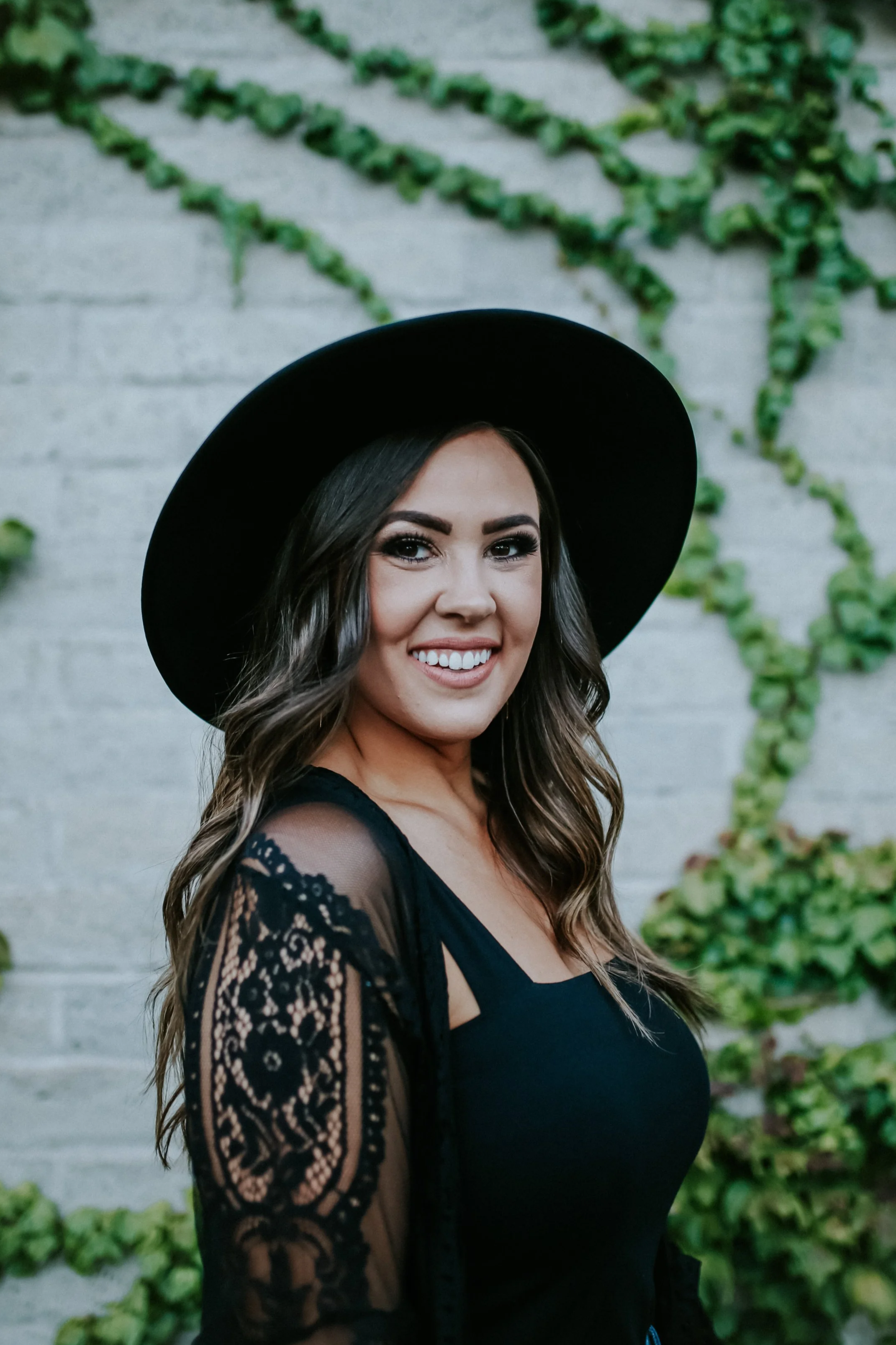 A woman with long wavy brown hair wearing a black wide-brim hat and black lace top stands in front of a white brick wall with climbing green ivy, smiling at the camera.