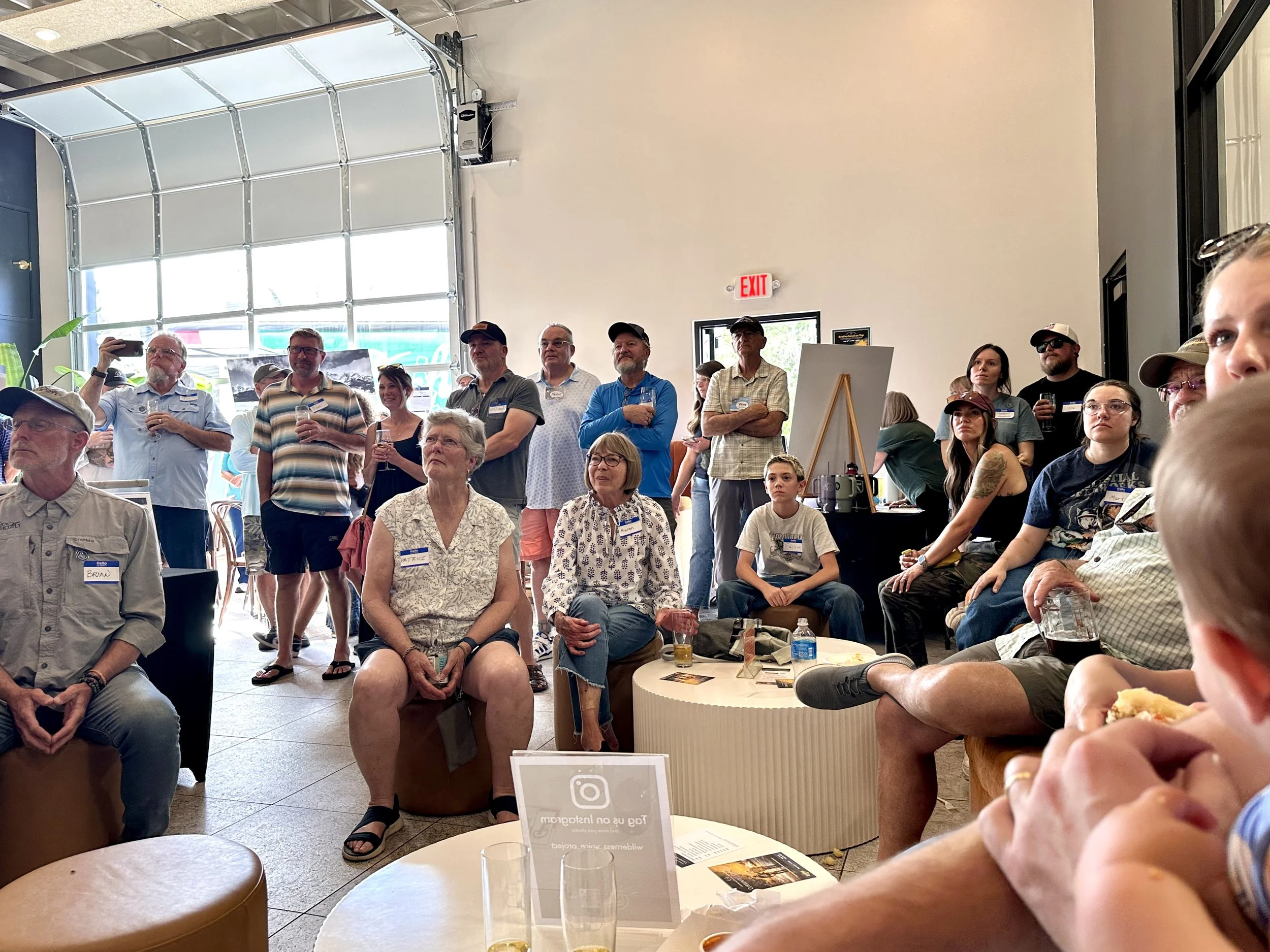 A group of people attending an indoor event or gathering, with some standing and others seated, in a spacious venue with large windows and a high ceiling.