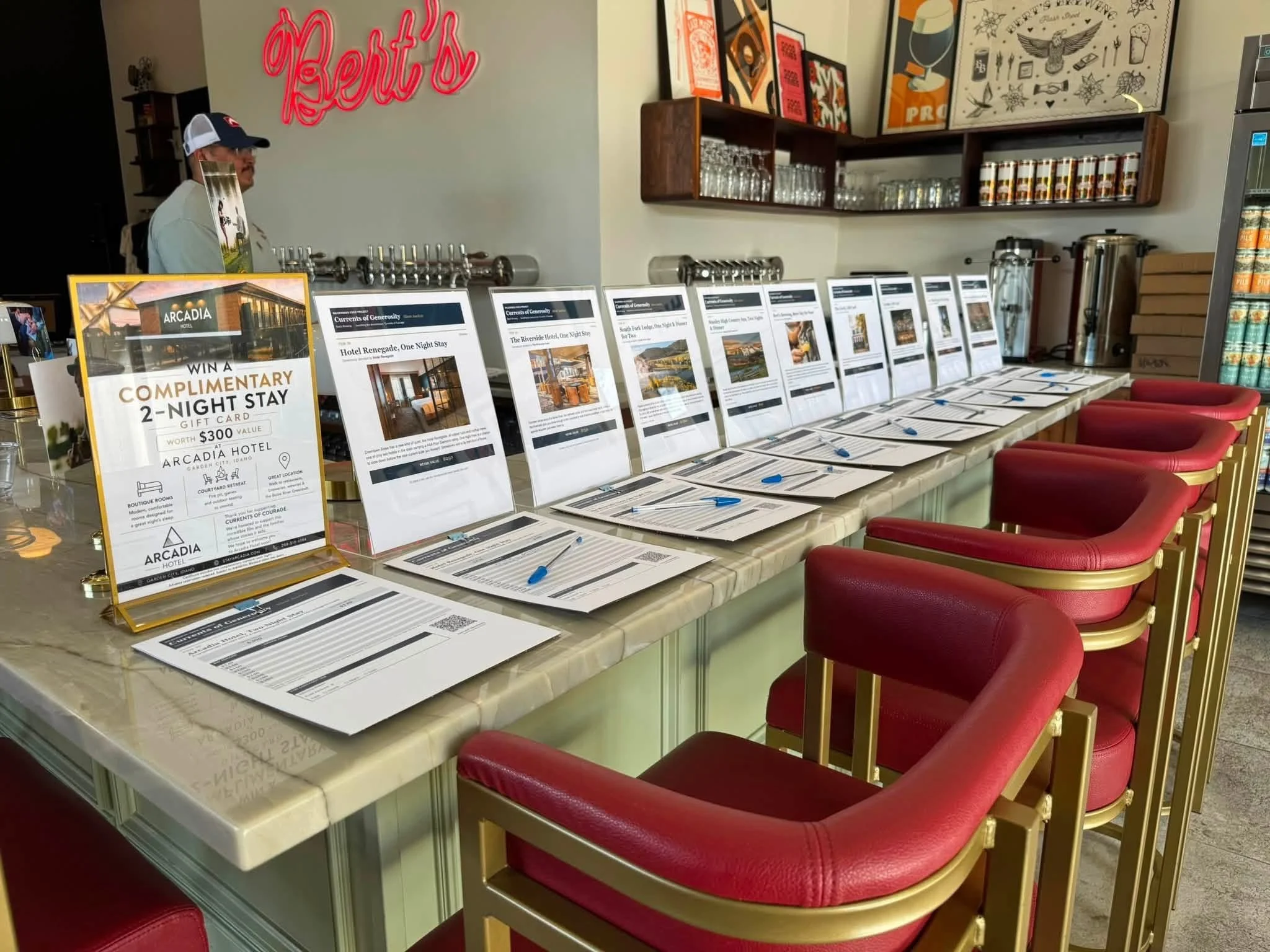 Display table with promotional flyers and brochures at a hotel reception, red chairs lined up along the front, shelves with glasses and beer taps, and colorful artwork on the walls.