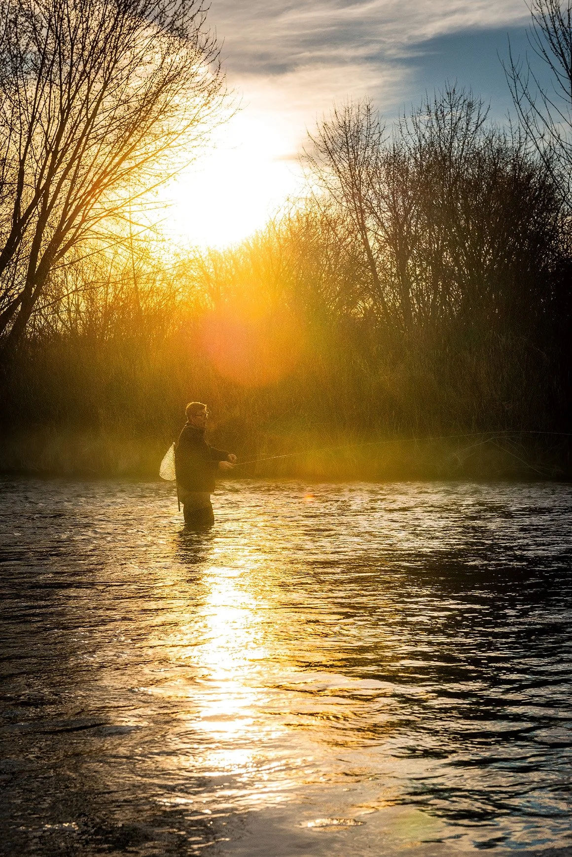 A person fishing in a river during sunset with bare trees in the background and sunlight reflecting on the water.