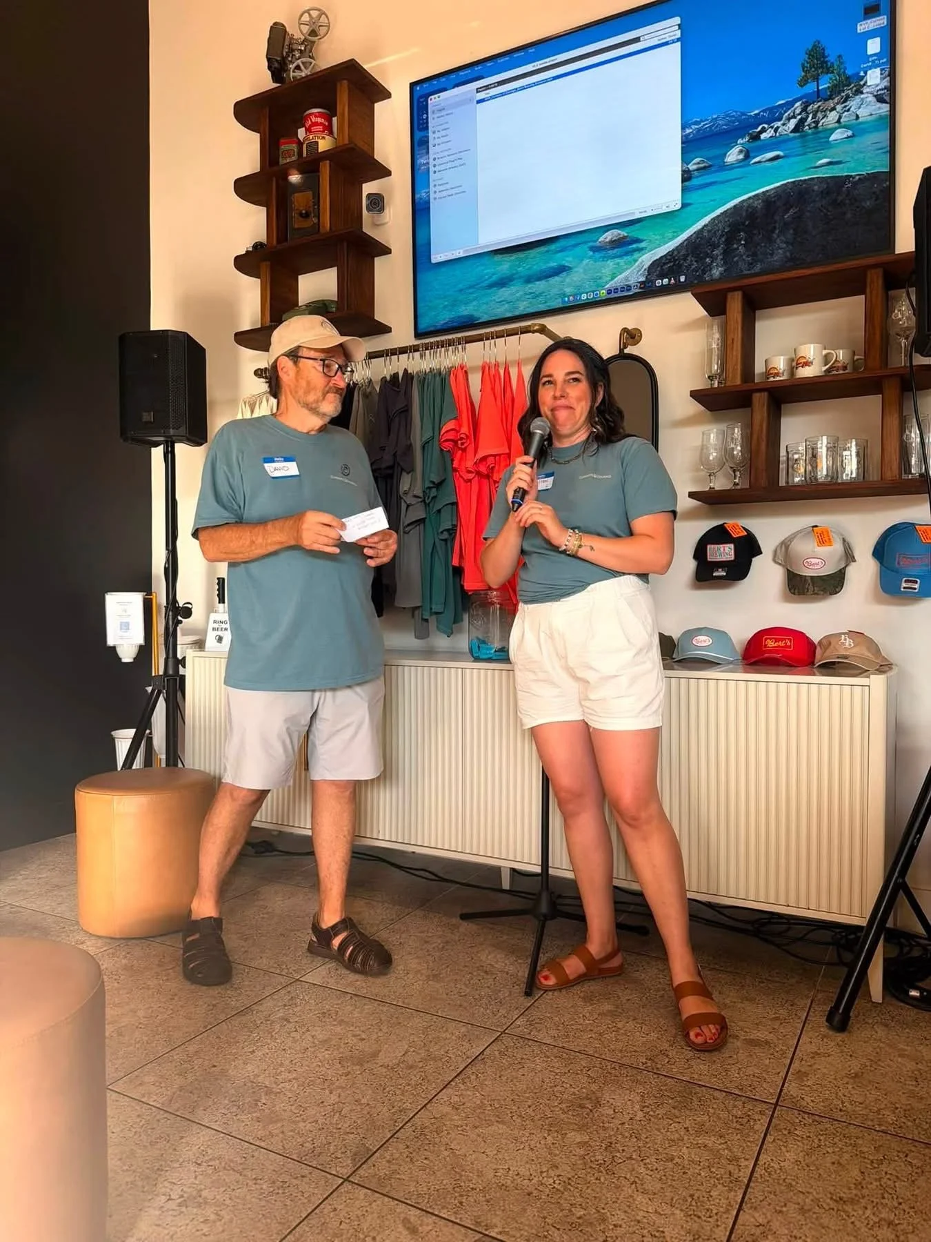 A man and woman standing indoors in front of a wall with shelves holding clothes, hats, and glassware. The woman is holding a microphone, and the man is holding a piece of paper. There is a large screen on the wall showing a scenic landscape of mountains, lake, and sky.