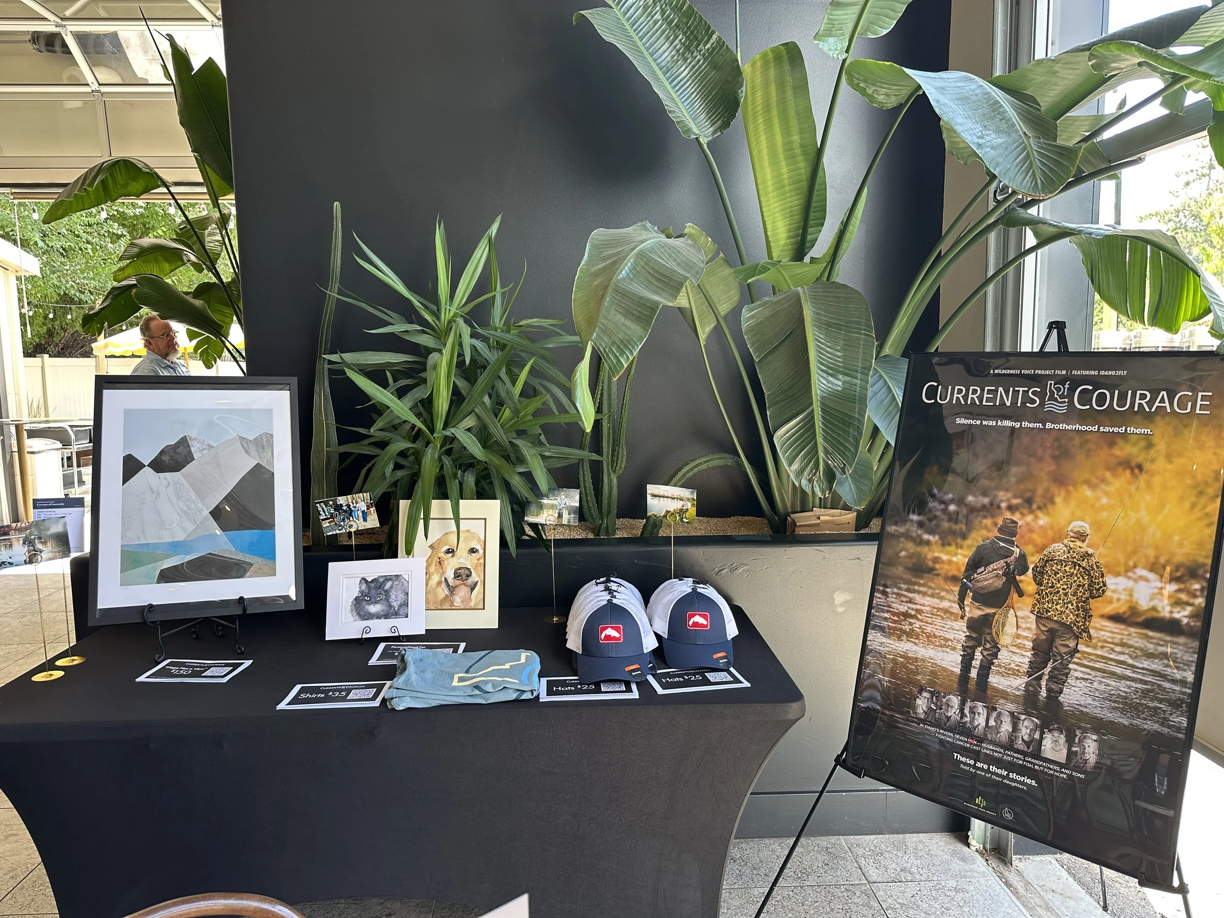 Display table with various merchandise including framed artwork, caps, t-shirts, and small photographs, with large houseplants in the background, inside a store or exhibit area.