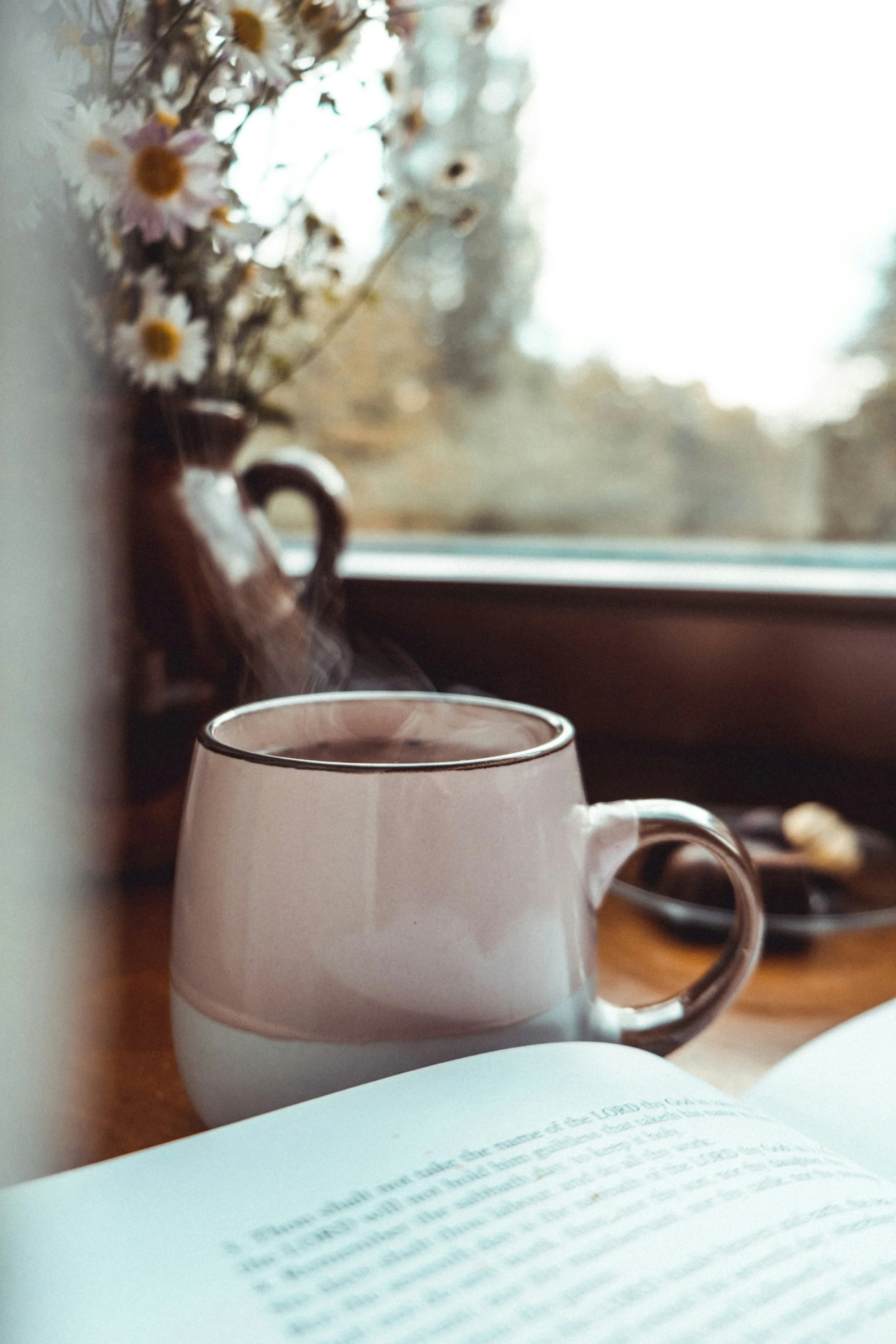 A steaming cup of tea on a wooden surface near a window, with a vase of daisies and an open book in the foreground.