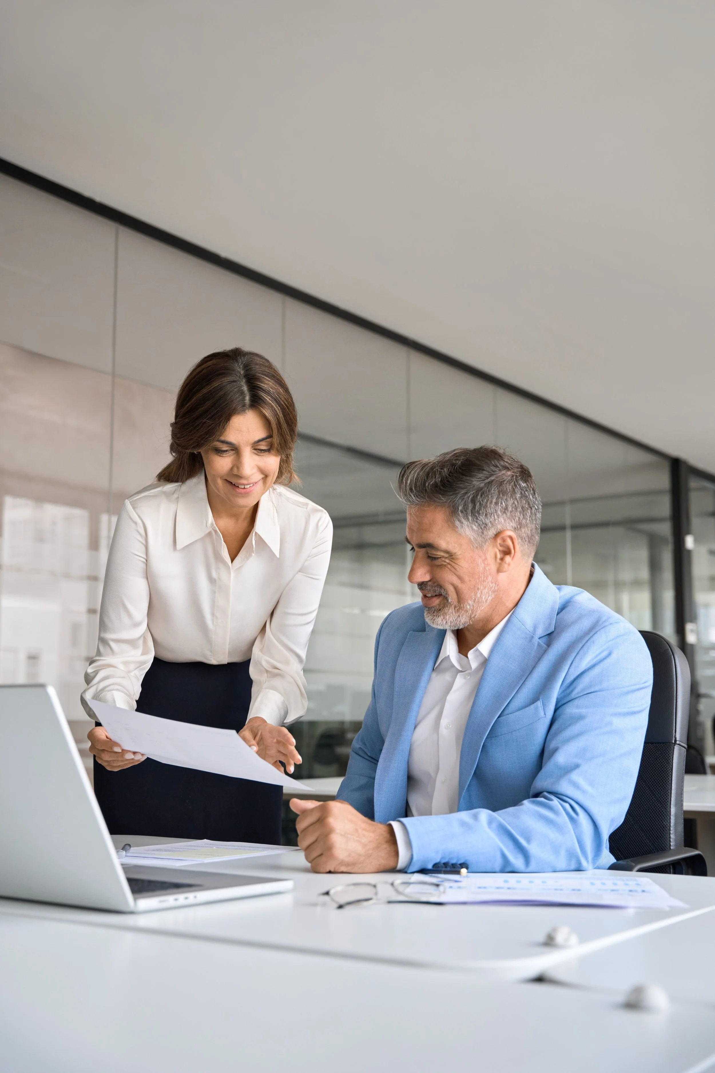 A woman and a man are having a discussion in an office, looking at documents and a laptop on the desk.