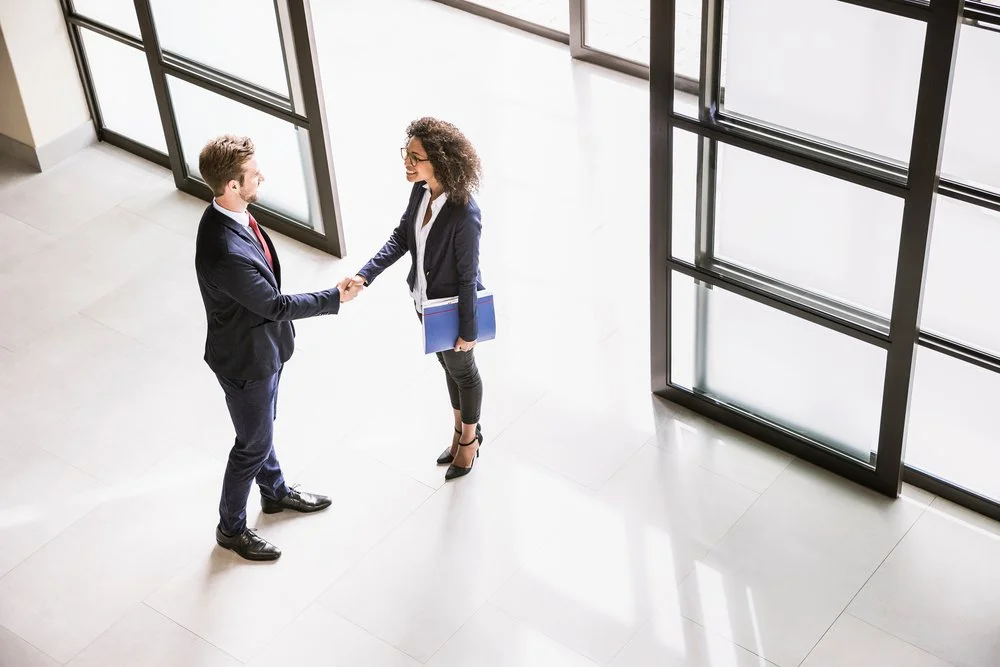 Two business professionals, a man and a woman, shaking hands in a modern office lobby with large windows.