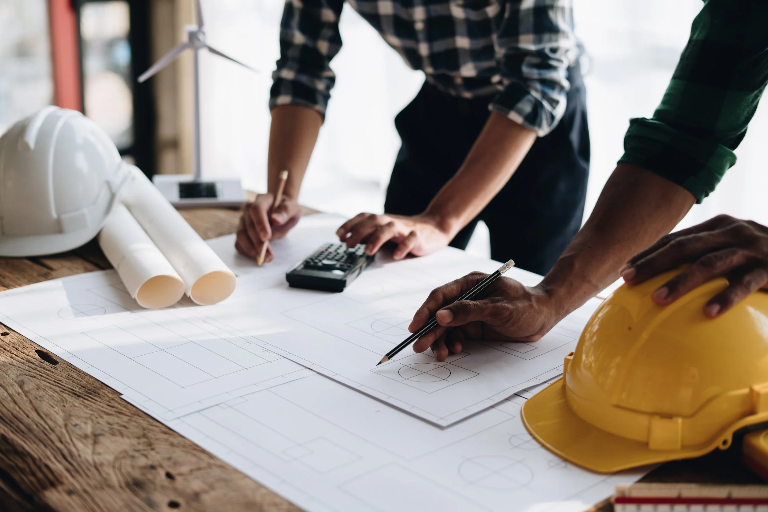 Two people working on building blueprints on a wooden table, with yellow and white construction helmets and rolled-up blueprints nearby.