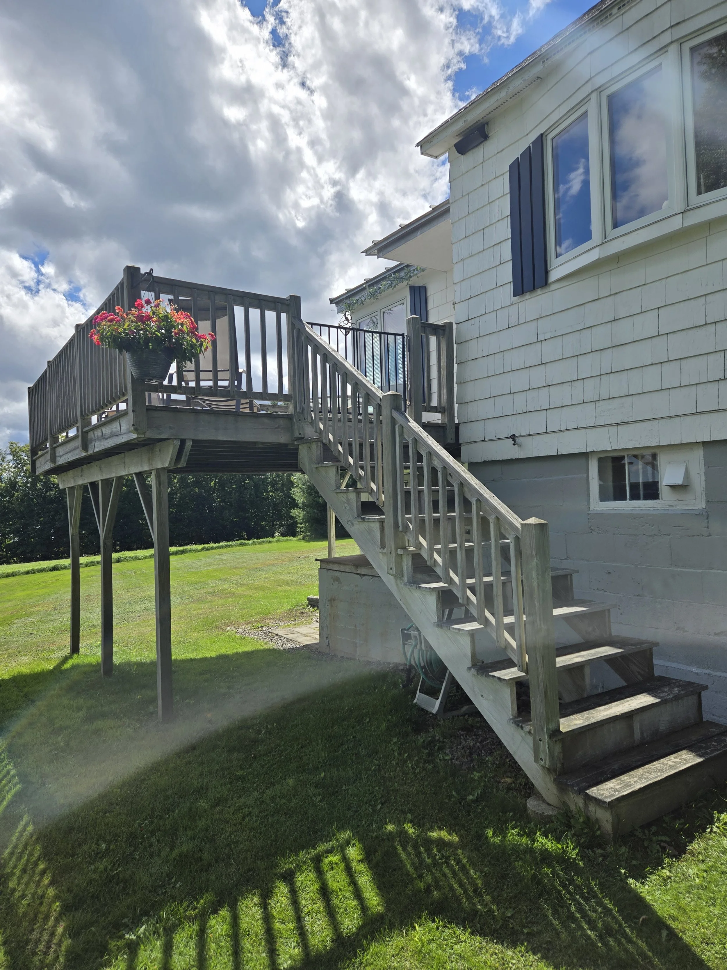 Wooden outdoor staircase leading to a house deck with potted flowers, on a grassy lawn under partly cloudy sky.