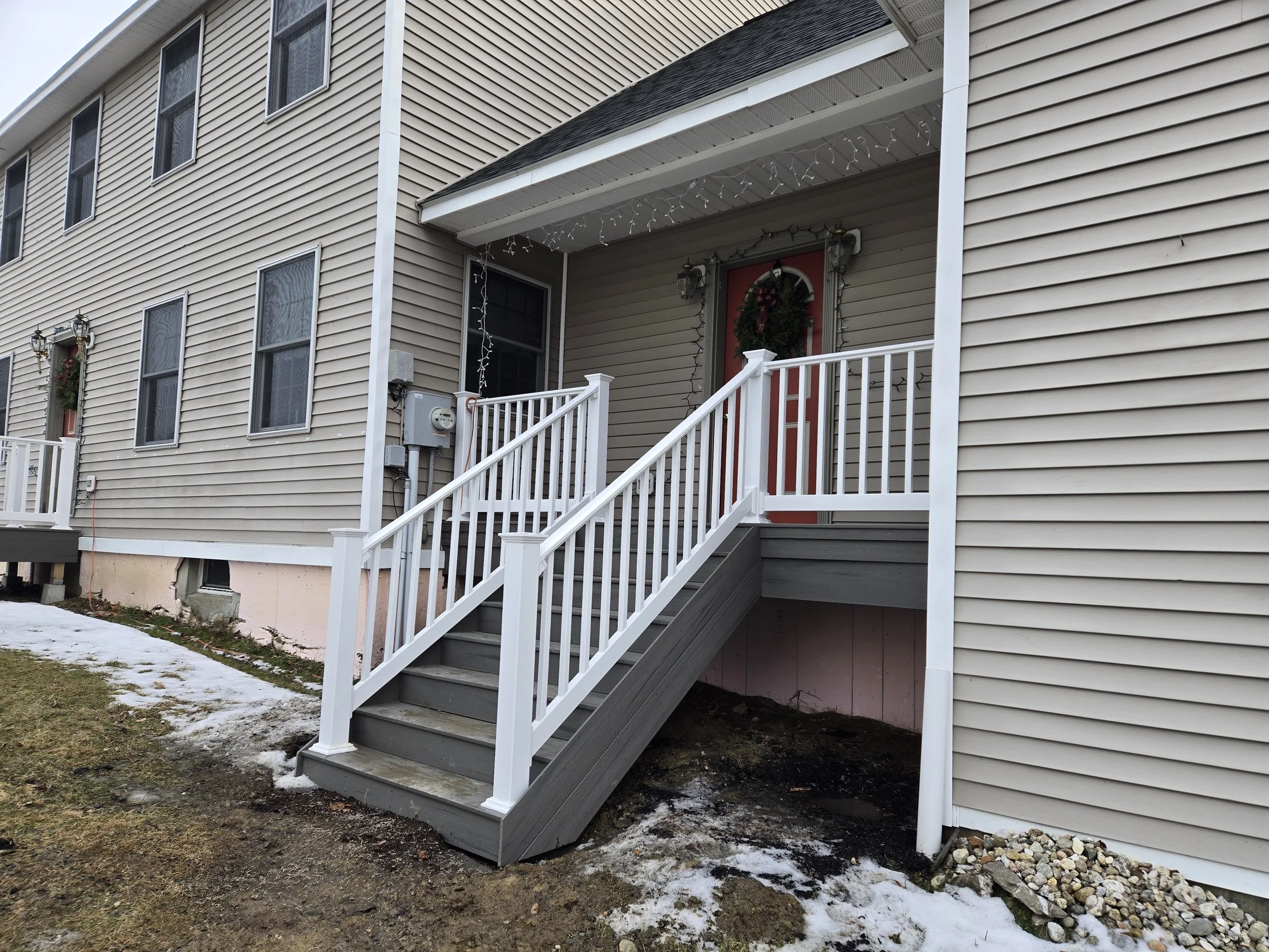 Front view of a beige townhouse with a small porch, white railing, steps, and a door decorated with a Christmas wreath. Snow covers part of the ground.